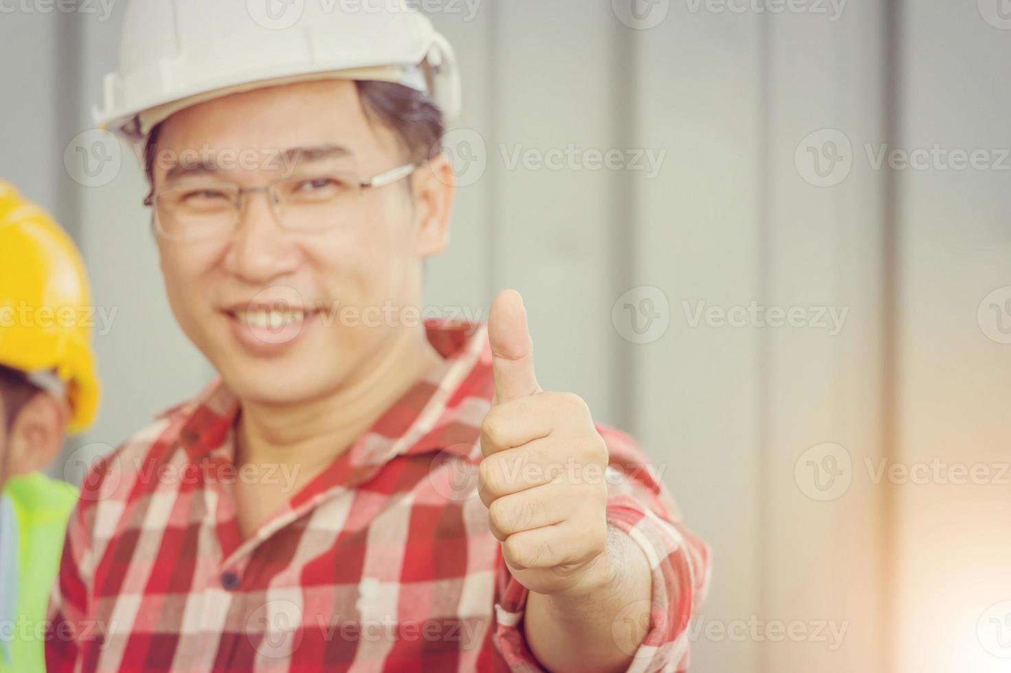 primo piano di un uomo sorridente dell'ingegnere aziendale che dà il pollice in su come segno di successo su un team di persone sfocate sullo sfondo del cantiere foto