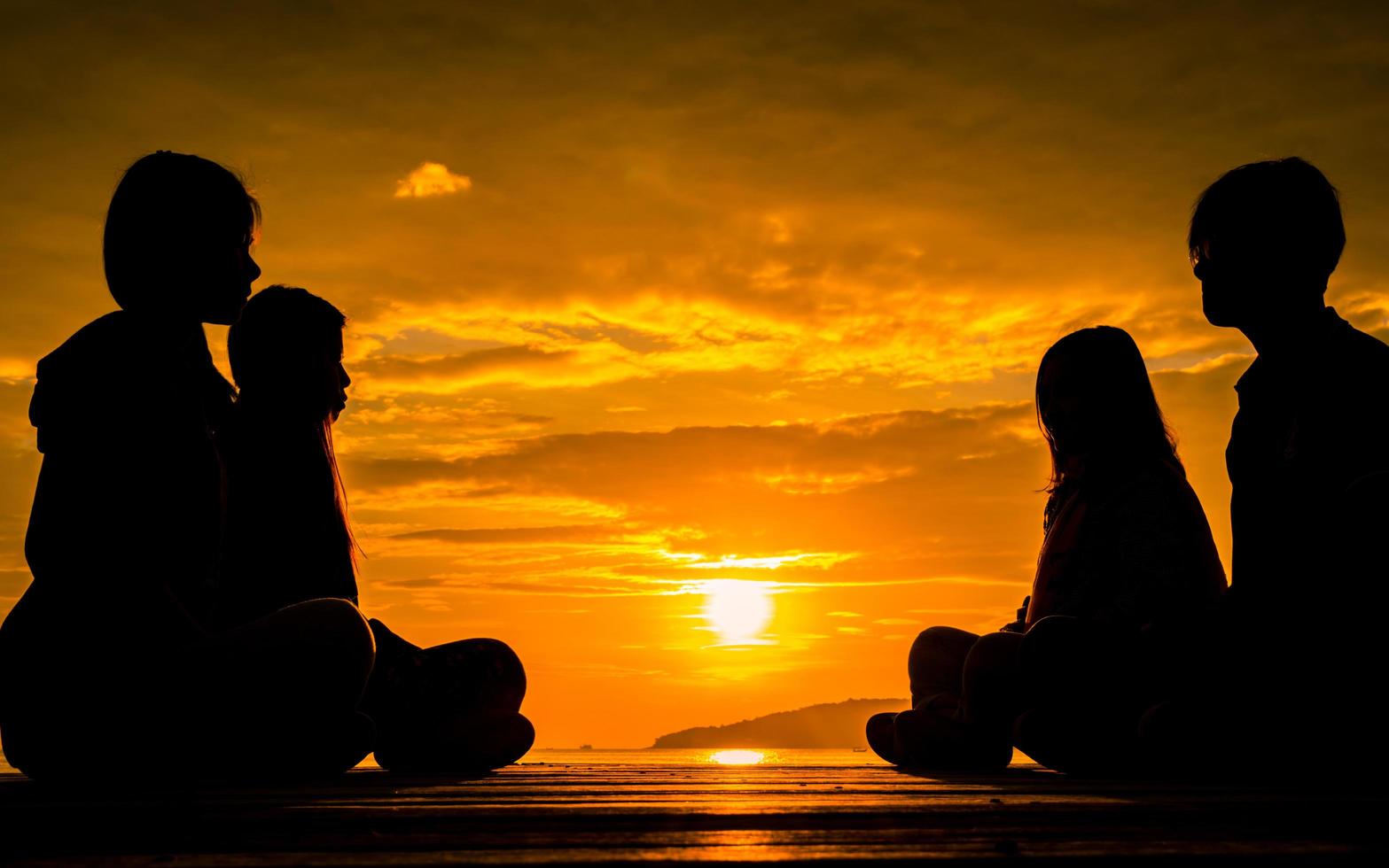 quattro giovani si siedono sul molo di legno all'alba sulla spiaggia per meditare con un bel cielo arancione e nuvole. foto