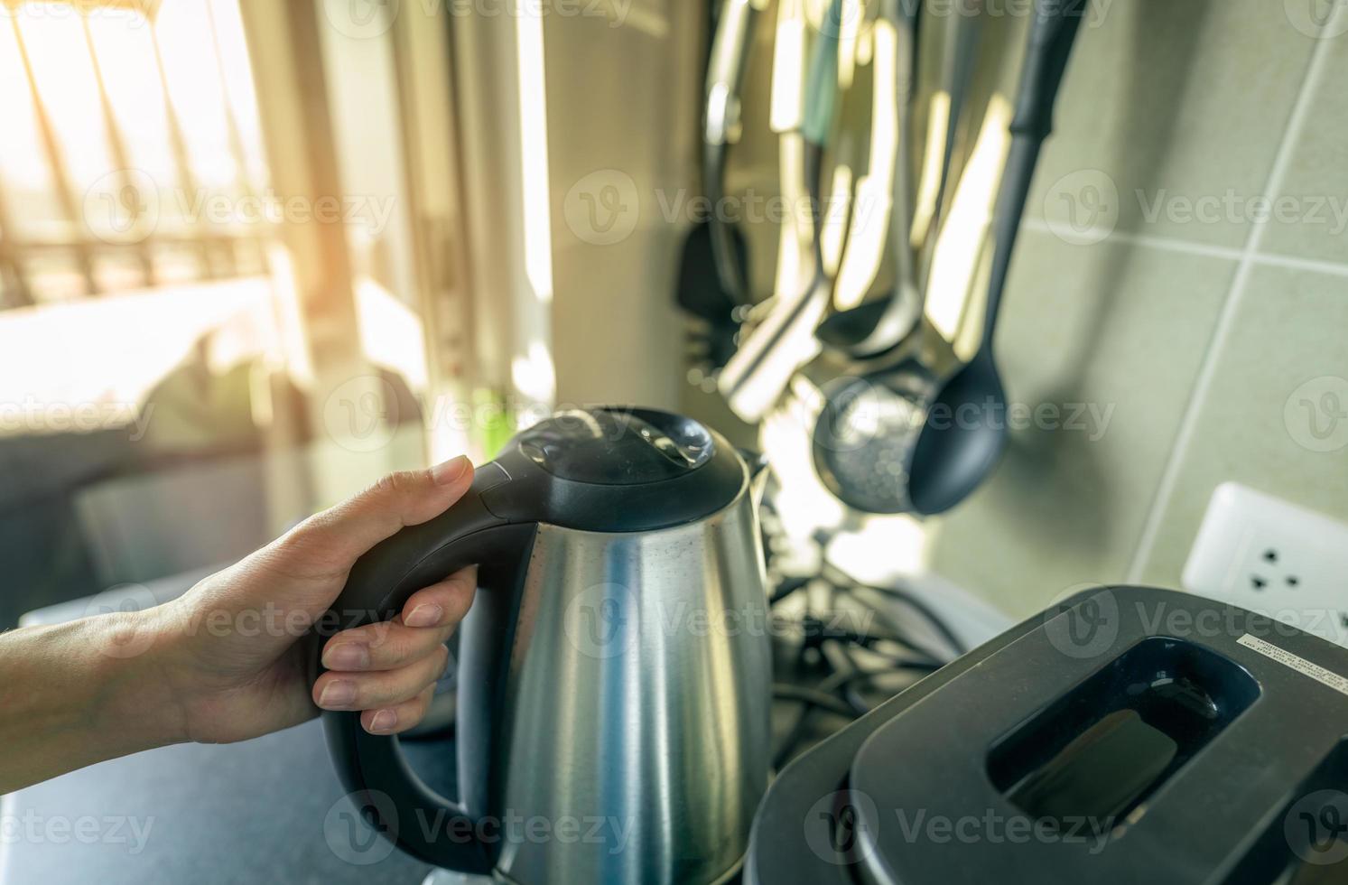 donna mano che tiene la maniglia del bollitore elettrico nella cucina dell'appartamento. la mano asiatica della donna prepara l'acqua bollita con il bollitore dell'acciaio inossidabile d'argento al mattino per la colazione, tè o caffè. elettrodomestico da cucina. foto