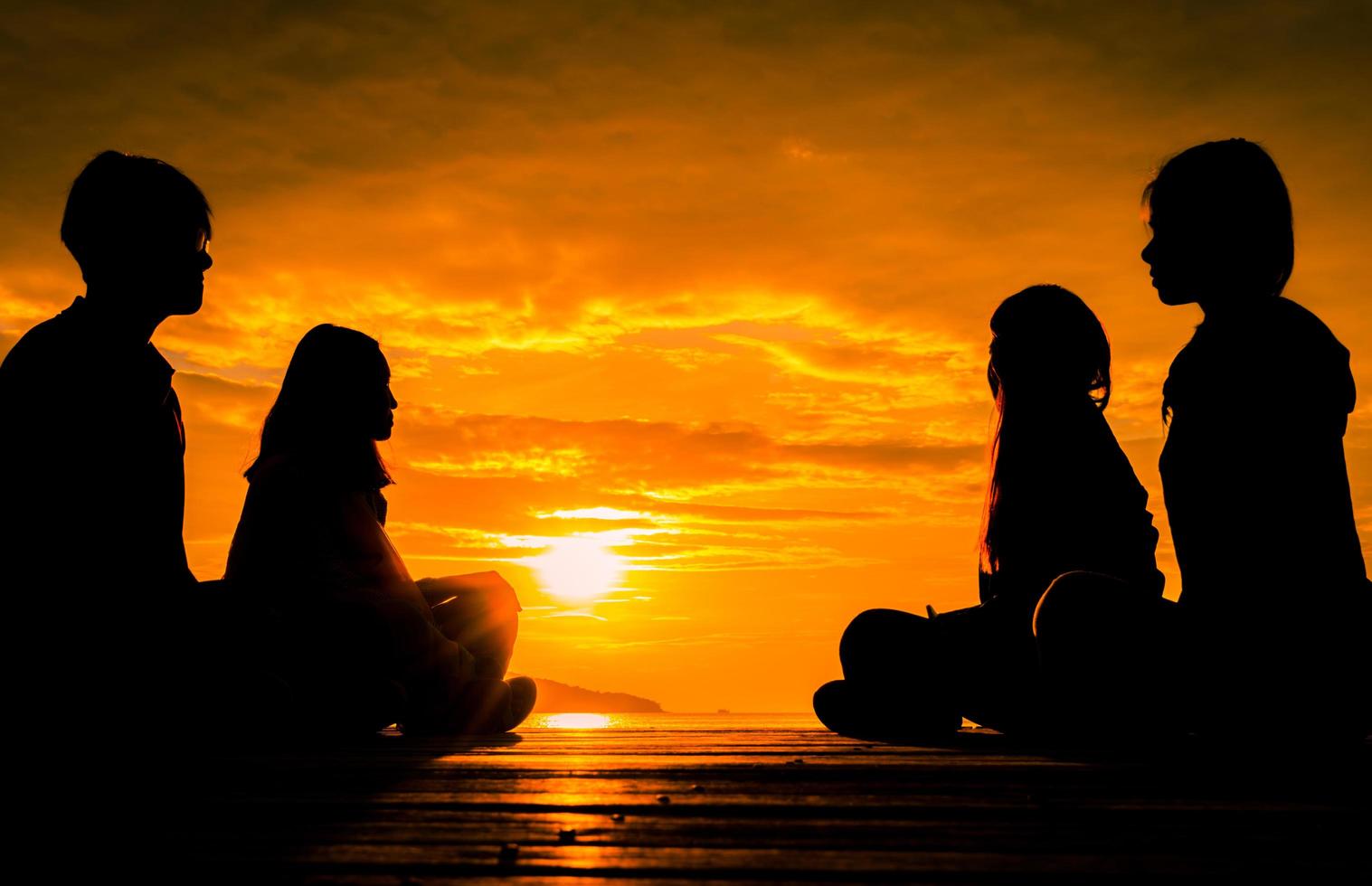 quattro giovani si siedono sul molo di legno all'alba sulla spiaggia per meditare con un bel cielo arancione e nuvole. foto