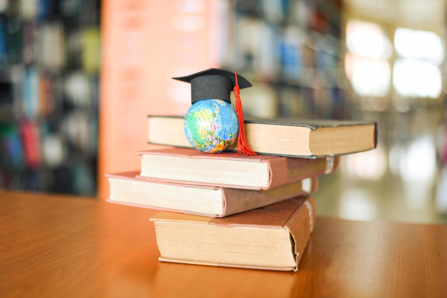 libri sul tavolo in biblioteca - istruzione che impara la pila di libri vecchi e il cappuccio di laurea sul modello del globo terrestre sulla scrivania in legno e sullo sfondo sfocato della stanza dello scaffale foto