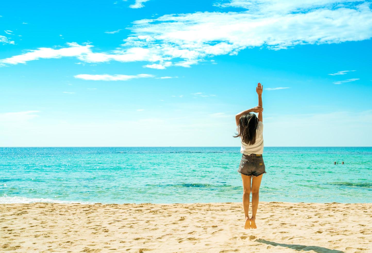 felice giovane donna in camicie bianche e pantaloncini che saltano sulla spiaggia di sabbia. rilassarsi e godersi le vacanze sulla spiaggia del paradiso tropicale con cielo blu e nuvole. ragazza in vacanza estiva. vibrazioni estive. giorno felice. foto