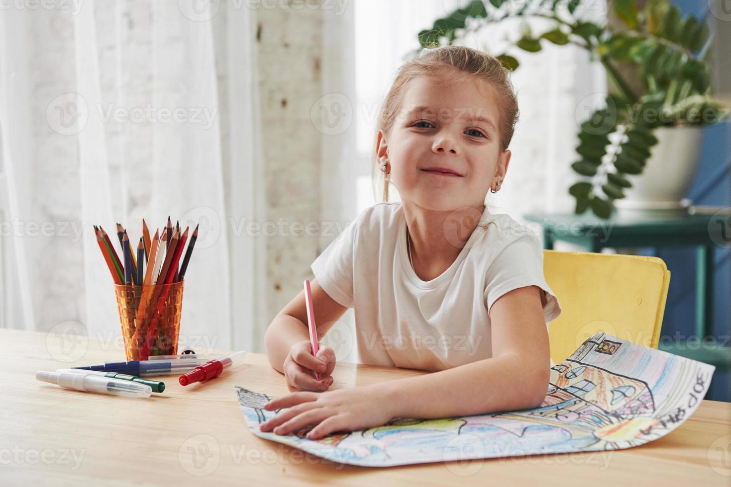 guardando nella telecamera con sguardo soddisfatto. la bambina carina della scuola d'arte disegna i suoi primi dipinti con matite e pennarelli foto