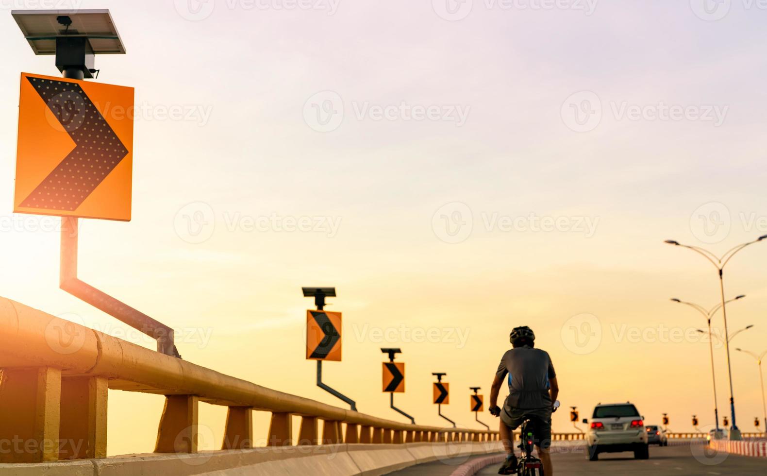 la vista posteriore dell'uomo indossa il casco in bicicletta sulla strada curva dietro le auto. guida sicura e guida su strada. segnale stradale di curva con energia del pannello solare. esercizio all'aperto. uno stile di vita sano. foto