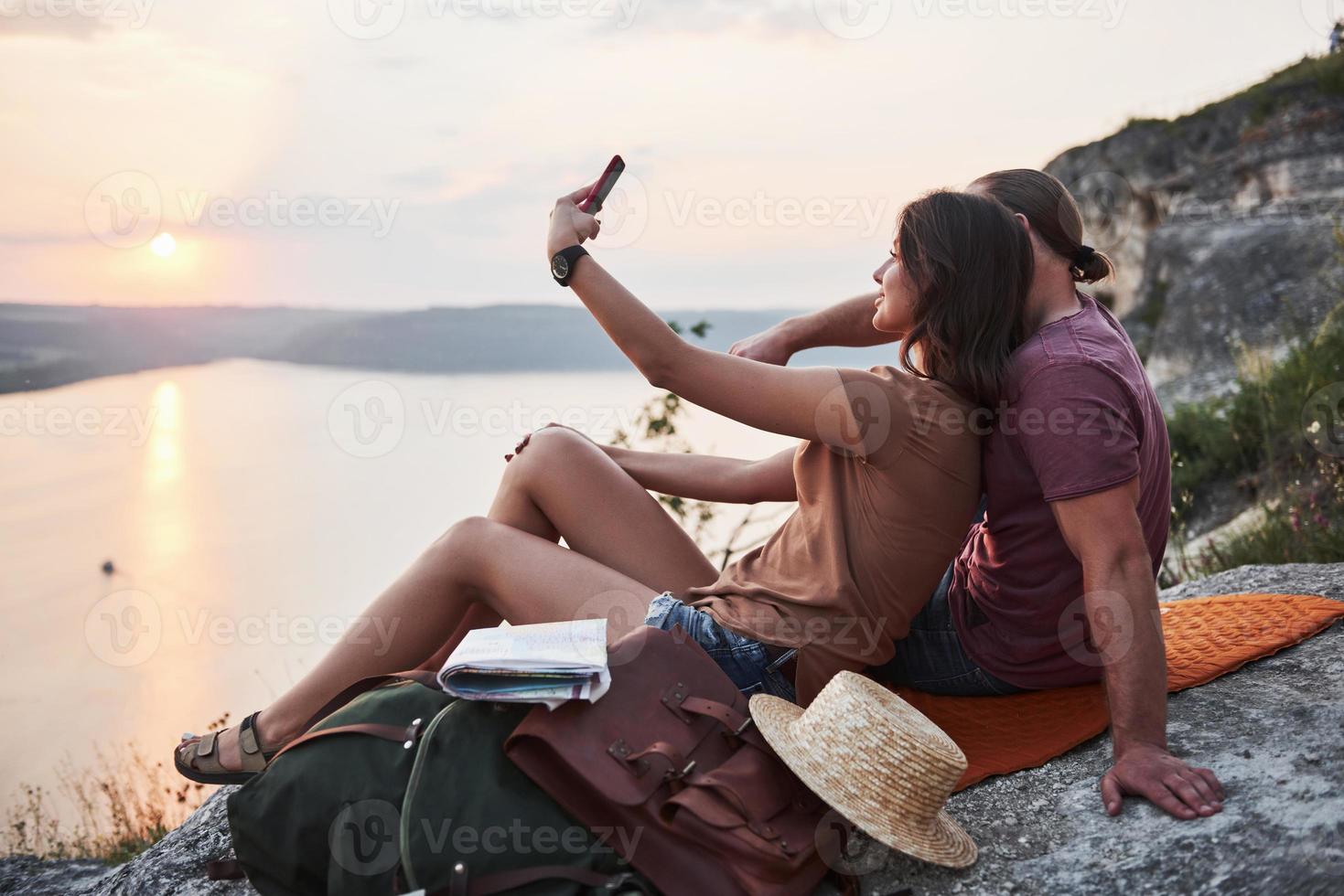 abbracciare la coppia con lo zaino seduto in cima alla montagna rocciosa godendo della vista sulla costa di un fiume o di un lago. viaggiando lungo le montagne e la costa, la libertà e il concetto di stile di vita attivo foto