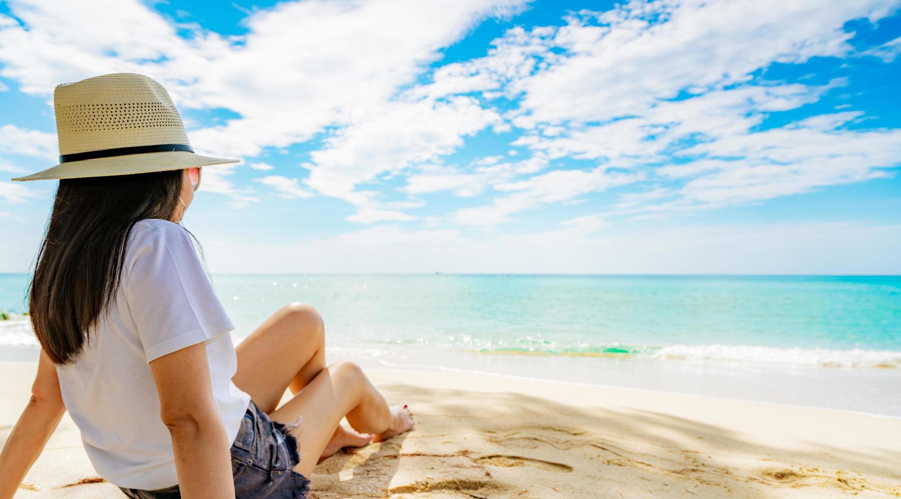 felice giovane donna in camicie bianche e pantaloncini seduti sulla spiaggia di sabbia. rilassarsi e godersi le vacanze sulla spiaggia del paradiso tropicale con cielo blu e nuvole. ragazza in vacanza estiva. vibrazioni estive. giorno felice. foto