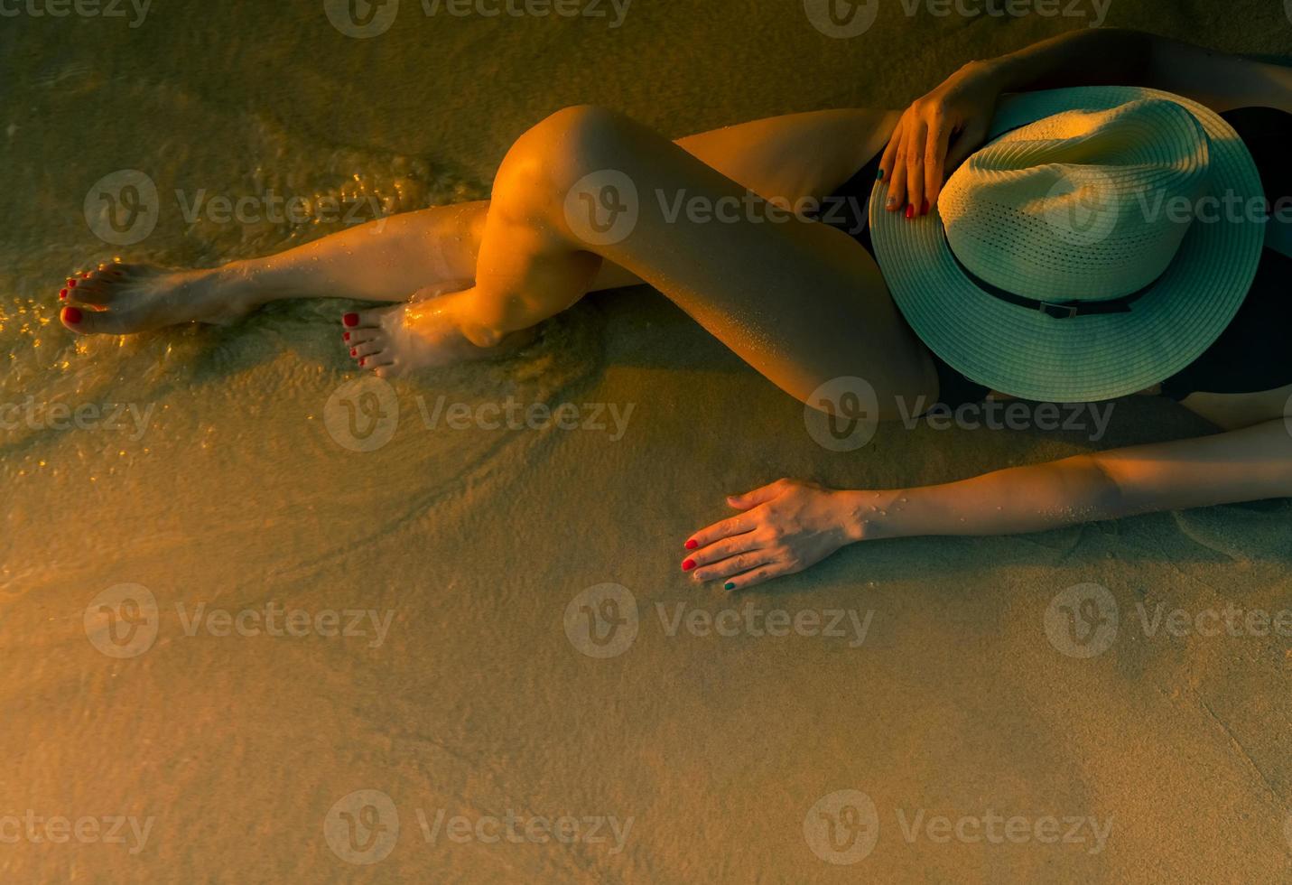 donna asiatica sexy con il cappello sdraiato sulla spiaggia di sabbia durante il tramonto. la donna indossa il costume da bagno nero che si rilassa sulla spiaggia. vacanze estive. estate. ragazza che prende il sole per la pelle abbronzata sulla spiaggia tropicale del paradiso foto