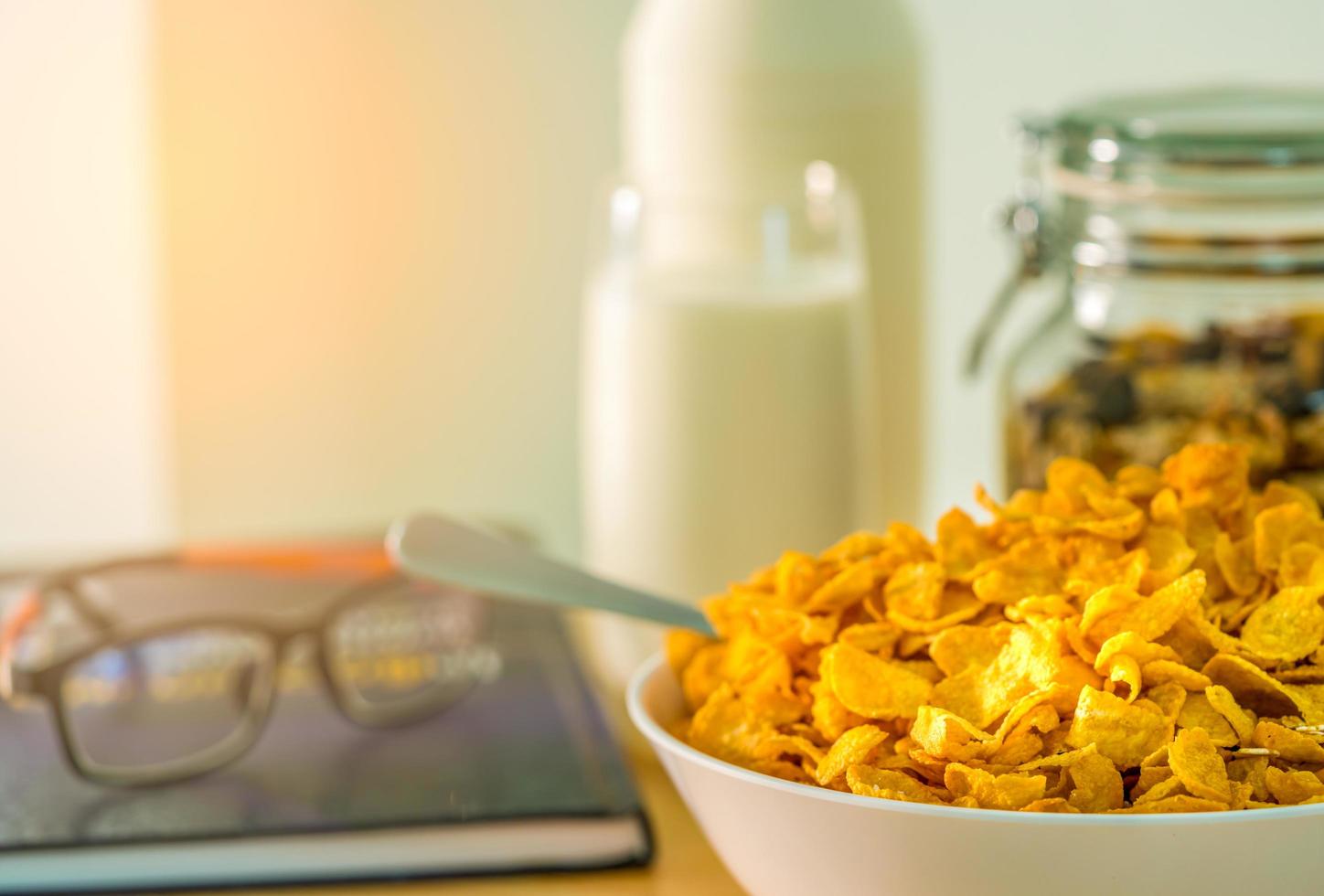 ciotola di cereali con cucchiaio sul tavolo di legno su sfondo sfocato di bicchiere di latte, bottiglia, contenitore di vetro, libro di testo e occhiali. cibo a base di calcio per colazione. cornflakes e concetto di latte. foto