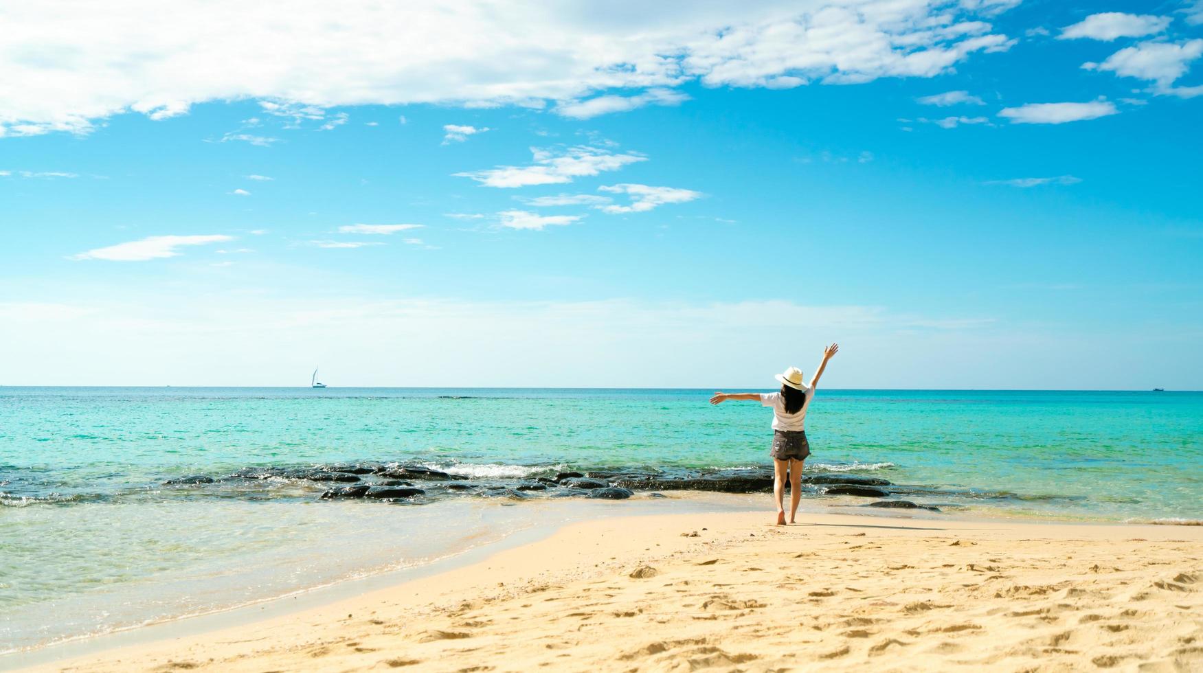 felice giovane donna in camicie bianche e pantaloncini che camminano sulla spiaggia di sabbia. rilassarsi e godersi le vacanze sulla spiaggia del paradiso tropicale con cielo blu e nuvole. ragazza in vacanza estiva. vibrazioni estive. giorno felice. foto