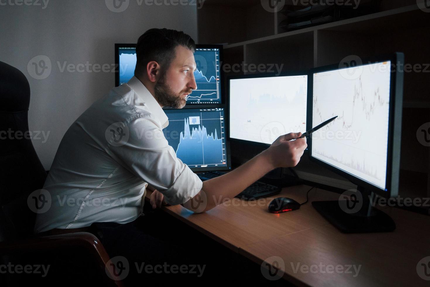 mostra il punto sul monitor dalla penna. uomo barbuto in camicia bianca lavora in ufficio con più schermi di computer nei grafici indice foto