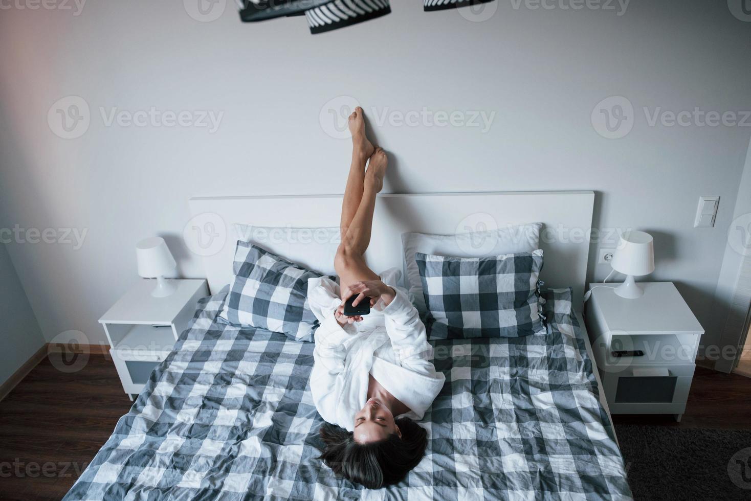 chiamando l'amico. bella ragazza in veste bianca sdraiata sul letto al mattino e utilizza lo smartphone foto