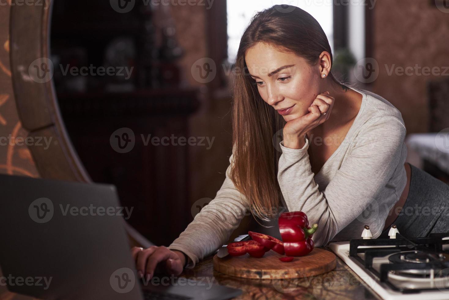 alla ricerca della ricetta bella giovane donna in piedi nella cucina moderna vicino al fornello a gas e utilizzando il laptop foto