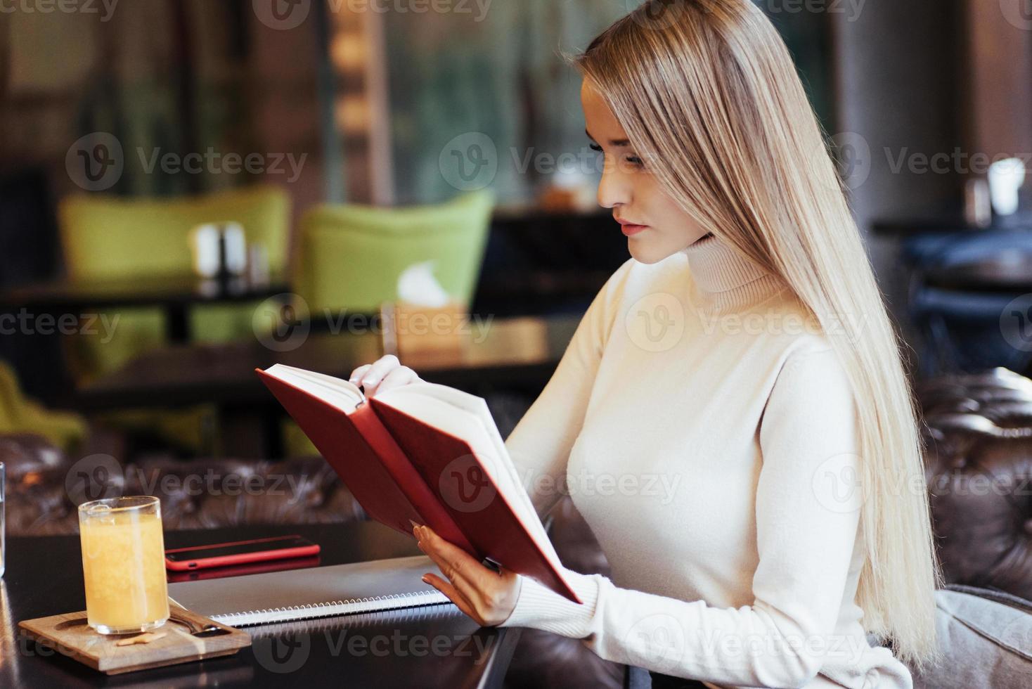 preparazione per gli esami. ragazza bionda ha letto un libro con la copertina rossa nel ristorante con un tavolo e una bevanda gialla su di esso foto