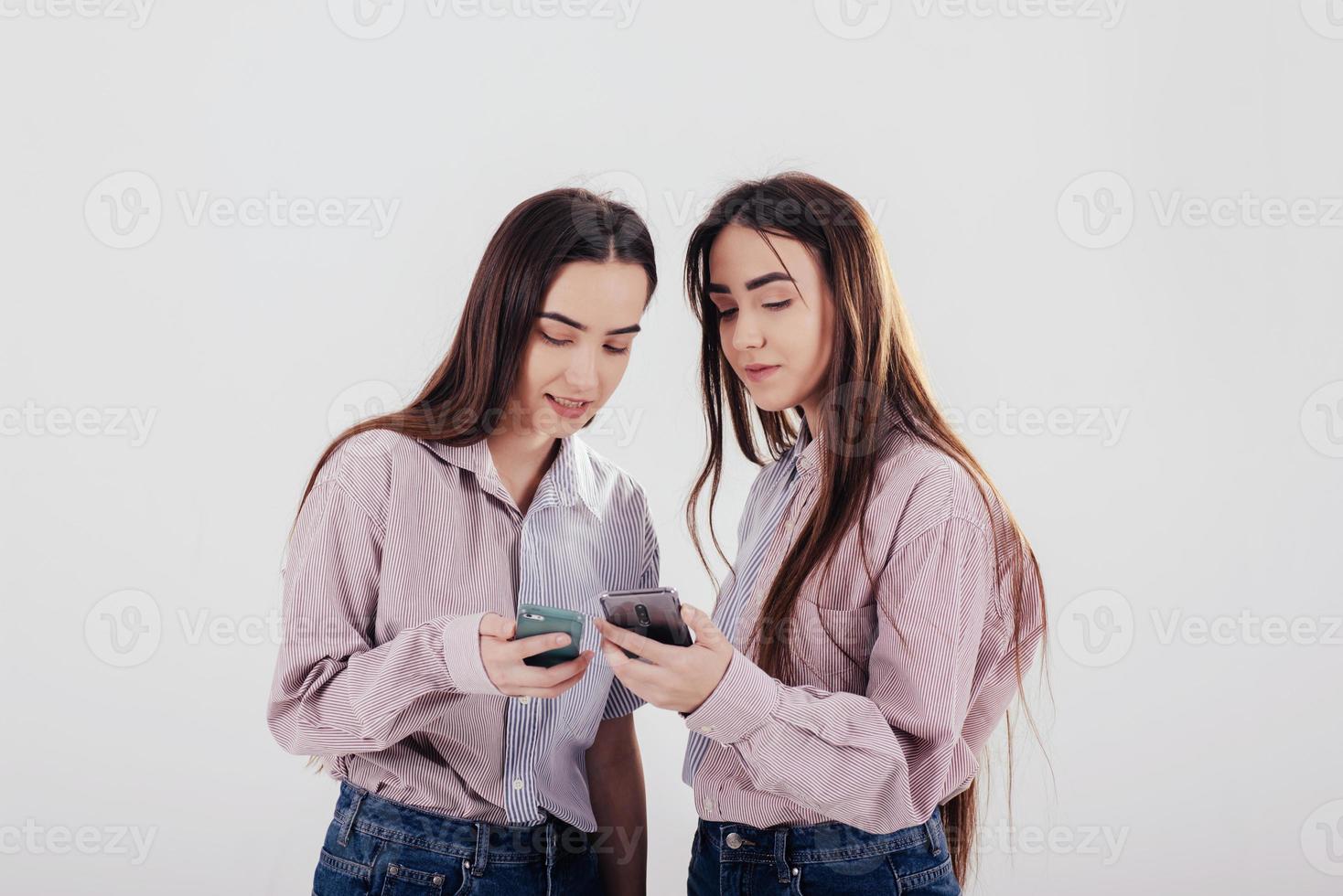 condivisione di alcune informazioni tramite smartphone. due sorelle gemelle in piedi e in posa in studio con sfondo bianco foto