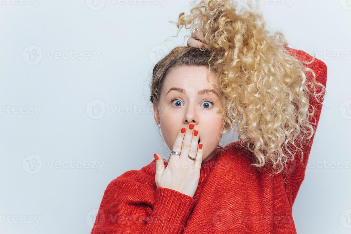 femmina bionda divertente emotiva con gli occhi infastiditi con coda di cavallo e capelli ricci, copre la bocca con la mano, non si aspetta di sentire notizie scioccanti da un amico, isolato su sfondo bianco. foto