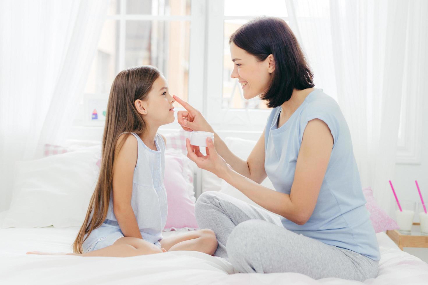 il ritratto di un bambino piccolo guarda felicemente la madre che si applica la crema sul naso, vestita con indumenti da notte, si sottopone a procedure di bellezza prima di dormire, si prende cura della propria pelle. concetto di famiglia, cosmetici e amore foto