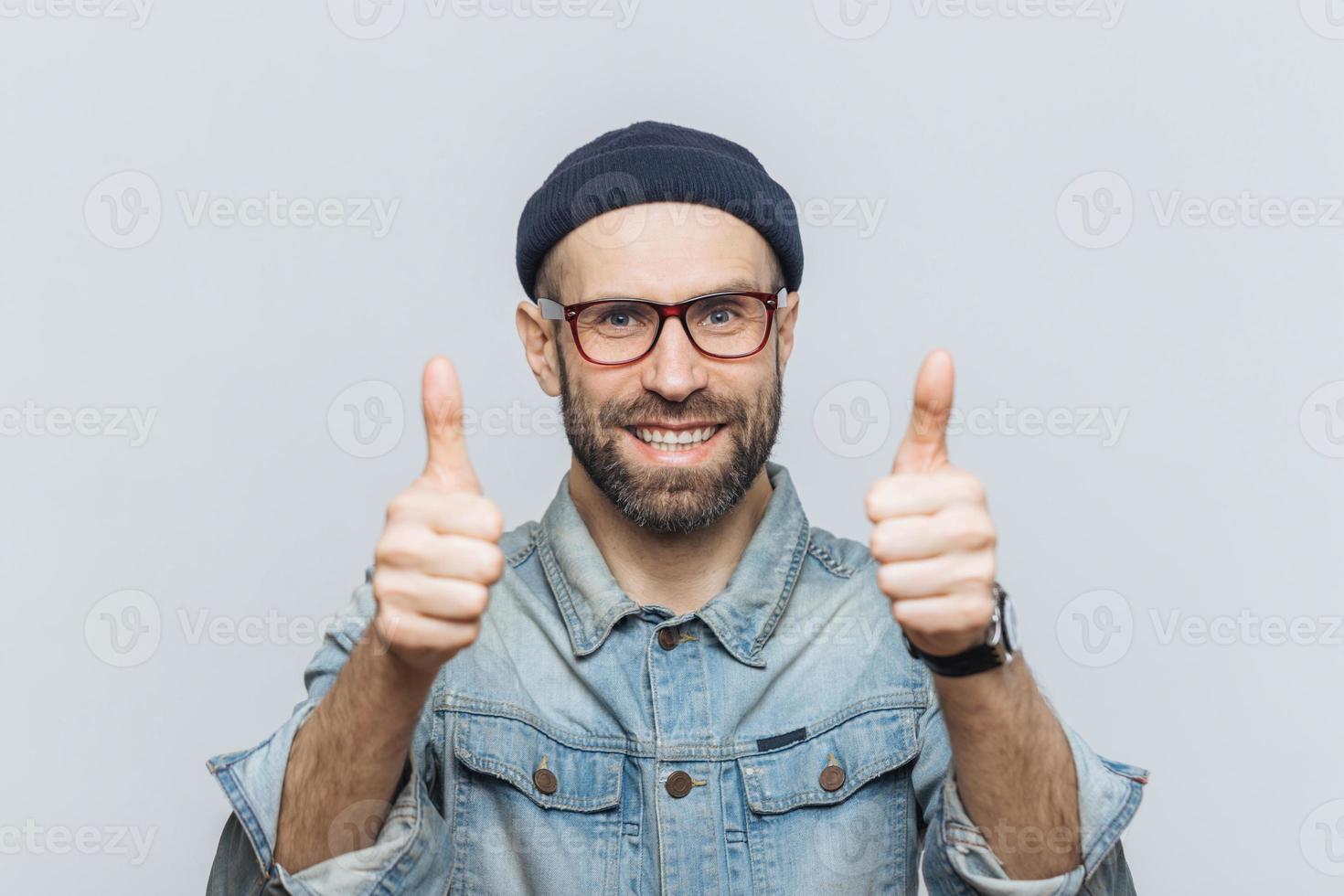 felice modello maschile barbuto indossa occhiali, cappello alla moda e giacca di jeans, tiene i pollici alzati, dimostra approvazione e accordo con qualcosa, ha un'espressione felice, isolato su muro grigio foto