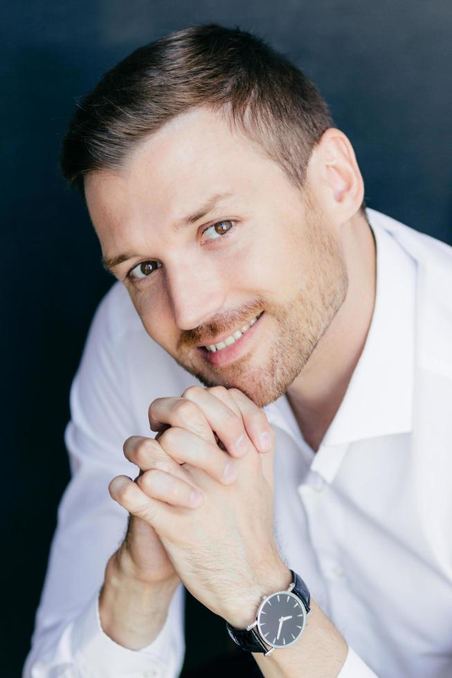 ritratto verticale di un bel maschio allegro con la barba lunga con un sorriso amichevole, tiene i palmi delle mani premuti insieme sotto il mento, indossa l'orologio a portata di mano, guarda felicemente la fotocamera. giovane lavoratore professionista foto