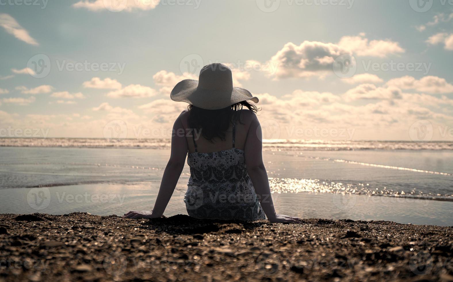 bella giovane donna ispanica seduta da sola sul bordo della spiaggia che indossa un cappello e un vestito bianco e nero durante il tramonto foto