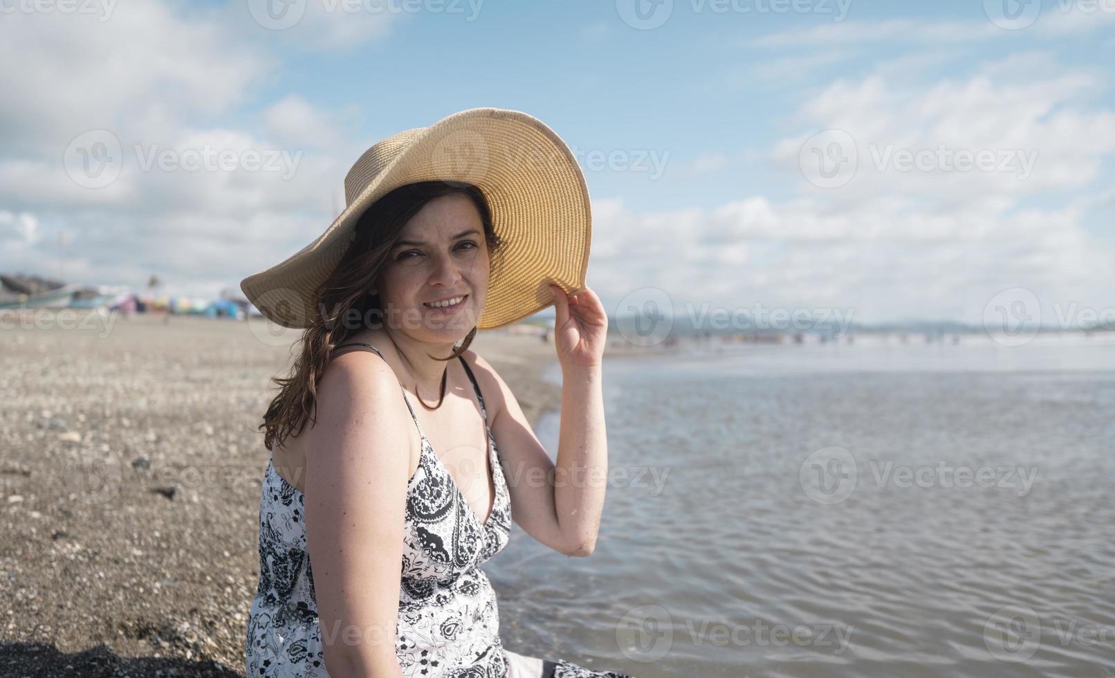 bella giovane donna ispanica felice seduta da sola sul bordo della spiaggia guardando la telecamera, indossando un cappello e un vestito bianco e nero durante una mattina di sole foto