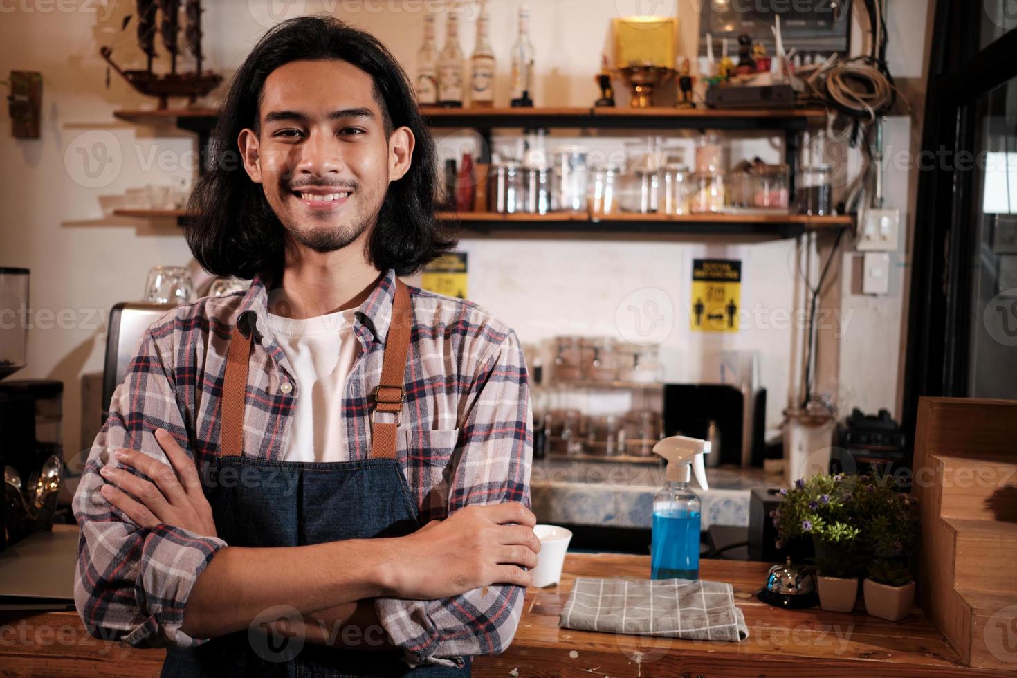 capelli lunghi bel barista asiatico di avvio maschile con grembiule si trova al bancone bar casual, braccia incrociate, guarda la fotocamera con un sorriso accogliente, felice e allegro con lavori di servizio di caffetteria. foto