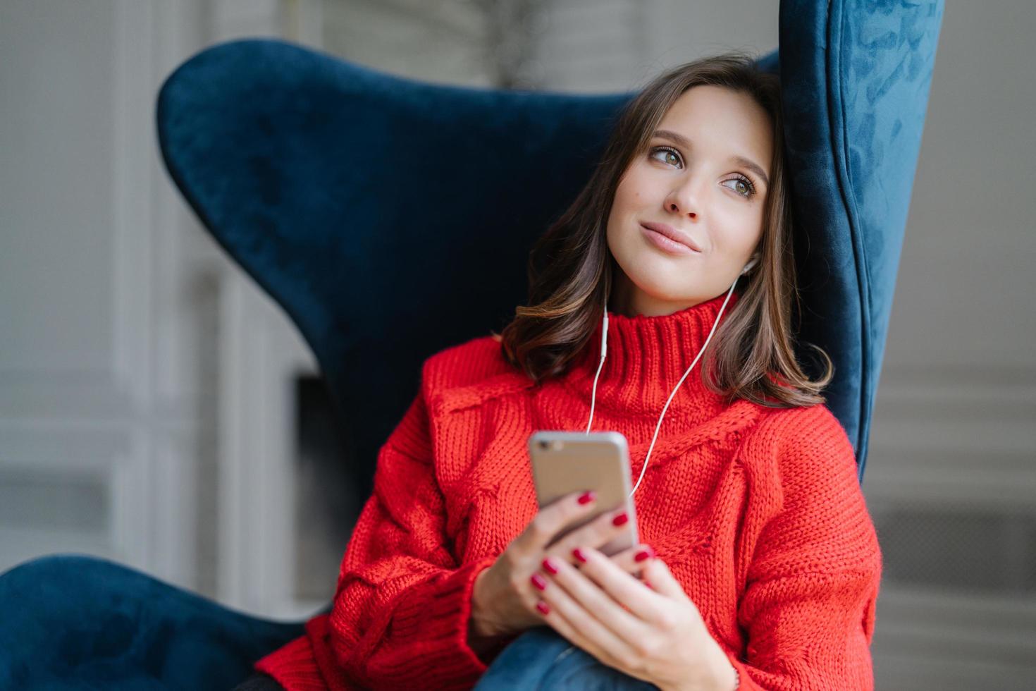 la donna carina e pensosa si sente rilassata, ascolta musica piacevole nella playlist, tiene il cellulare, guarda da parte con un'espressione sognante, posa in una comoda poltrona. persone, tecnologie moderne e intimità foto