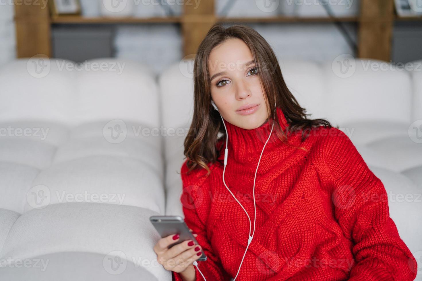 foto di una giovane modella rilassata che gode del tempo libero a casa, posa sul divano bianco, usa smartphone e auricolari, guarda film online, vestita con un maglione caldo lavorato a maglia, si gode il tempo a casa