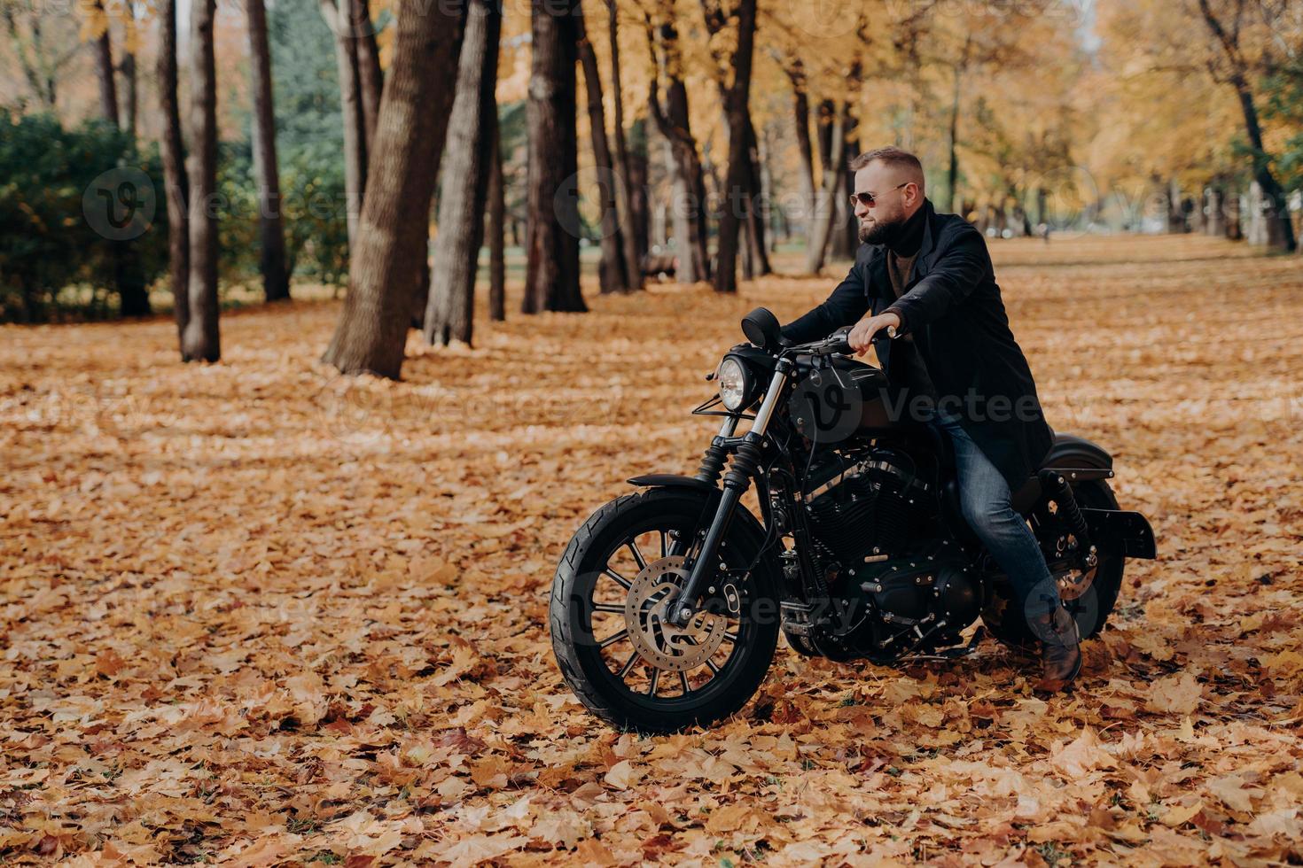 vista esterna orizzontale del motociclista maschio attivo va in bicicletta, indossa occhiali da sole alla moda e giacca nera, posa nel grande parco autunnale, gode di viaggi fantastici, fa un viaggio in moto, riposa nella natura. foto