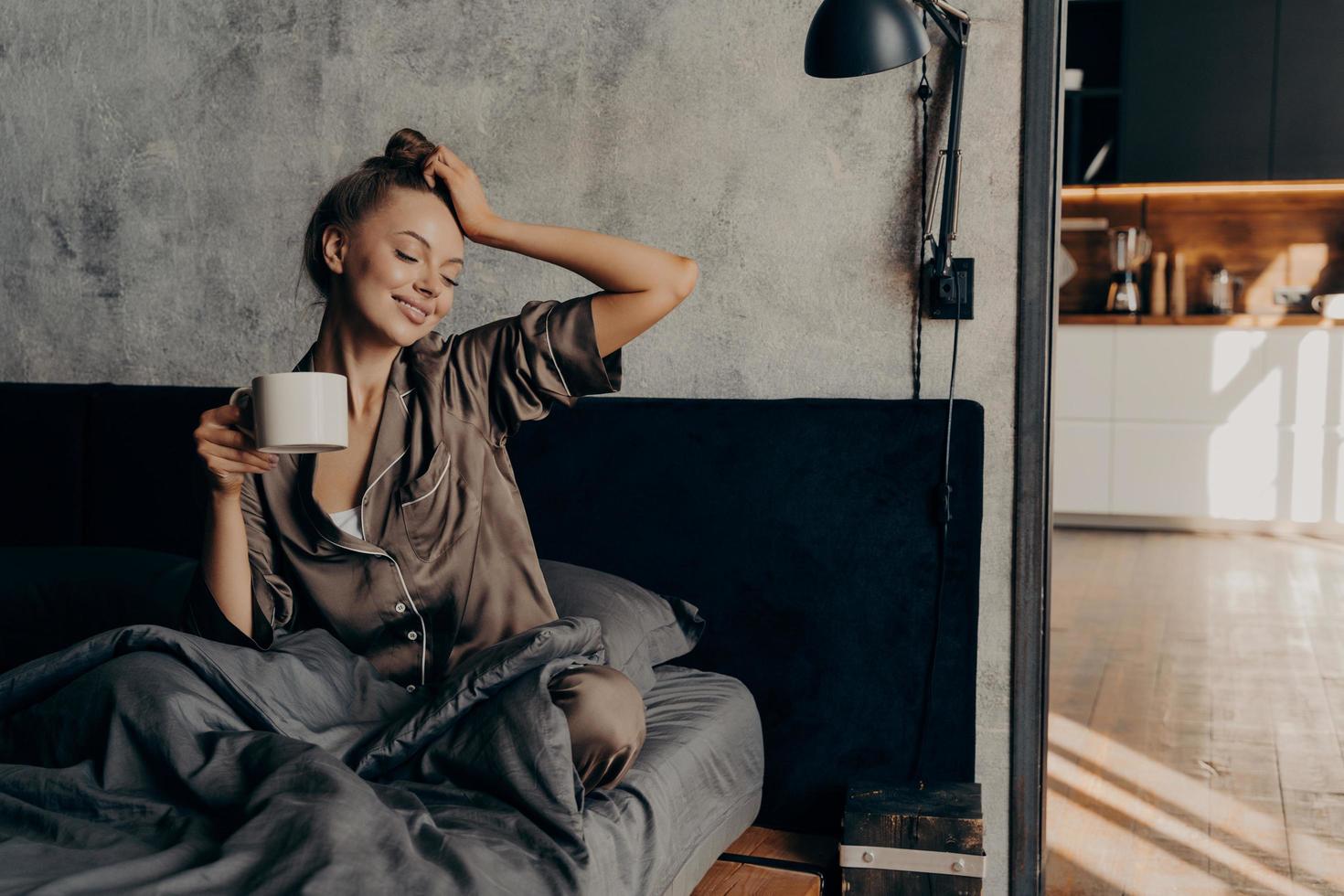 ritratto di giovane donna rilassata in un elegante abito da casa che si sveglia con una tazza di caffè a letto foto