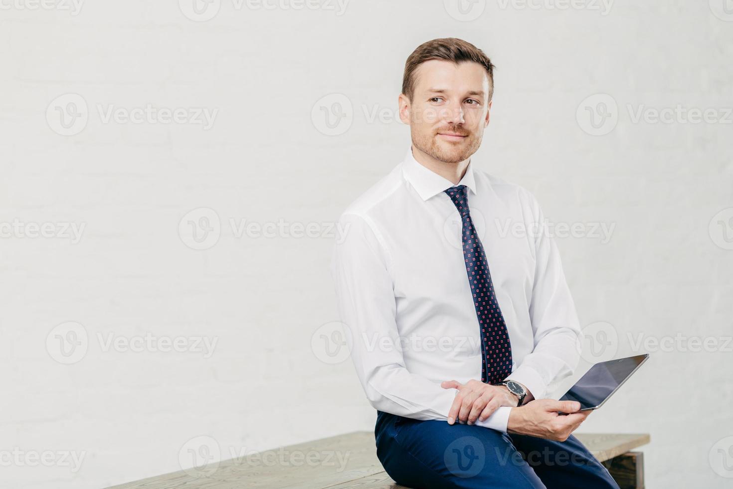 premuroso giovane economista maschio vestito con eleganti abiti di lusso, lavora sul touch pad durante la pausa, posa nel corridoio dell'ufficio moderno, pensa alla cooperazione, isolato su sfondo bianco. foto