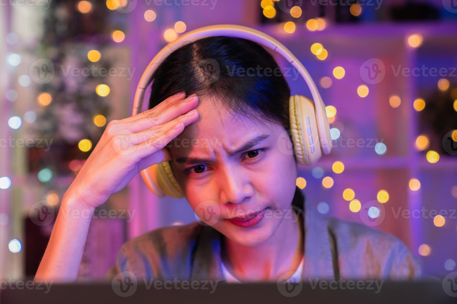 giovane donna asiatica stressante che indossa la cuffia e si siede sulla sedia con una pila di documenti sul tavolo e guardando lo schermo del computer di notte. foto