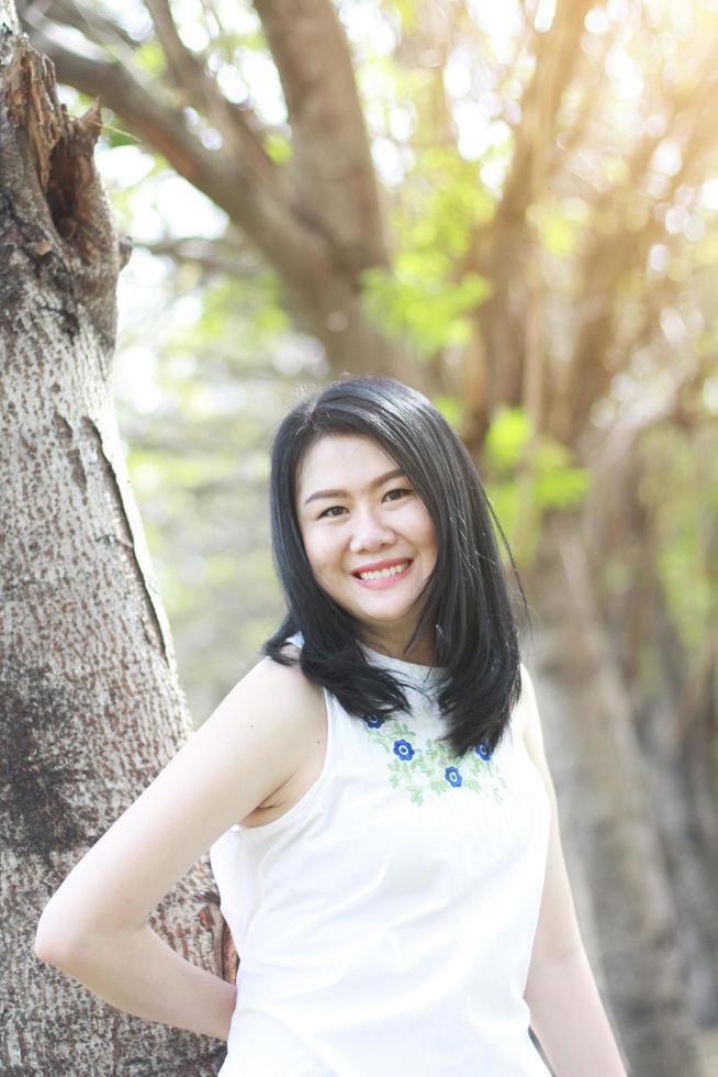 bella donna asiatica in abito bianco rilassarsi e sorridere nel parco naturale. la ragazza tailandese o la ragazza cinese si divertono in vacanza con la luce del sole in giardino foto
