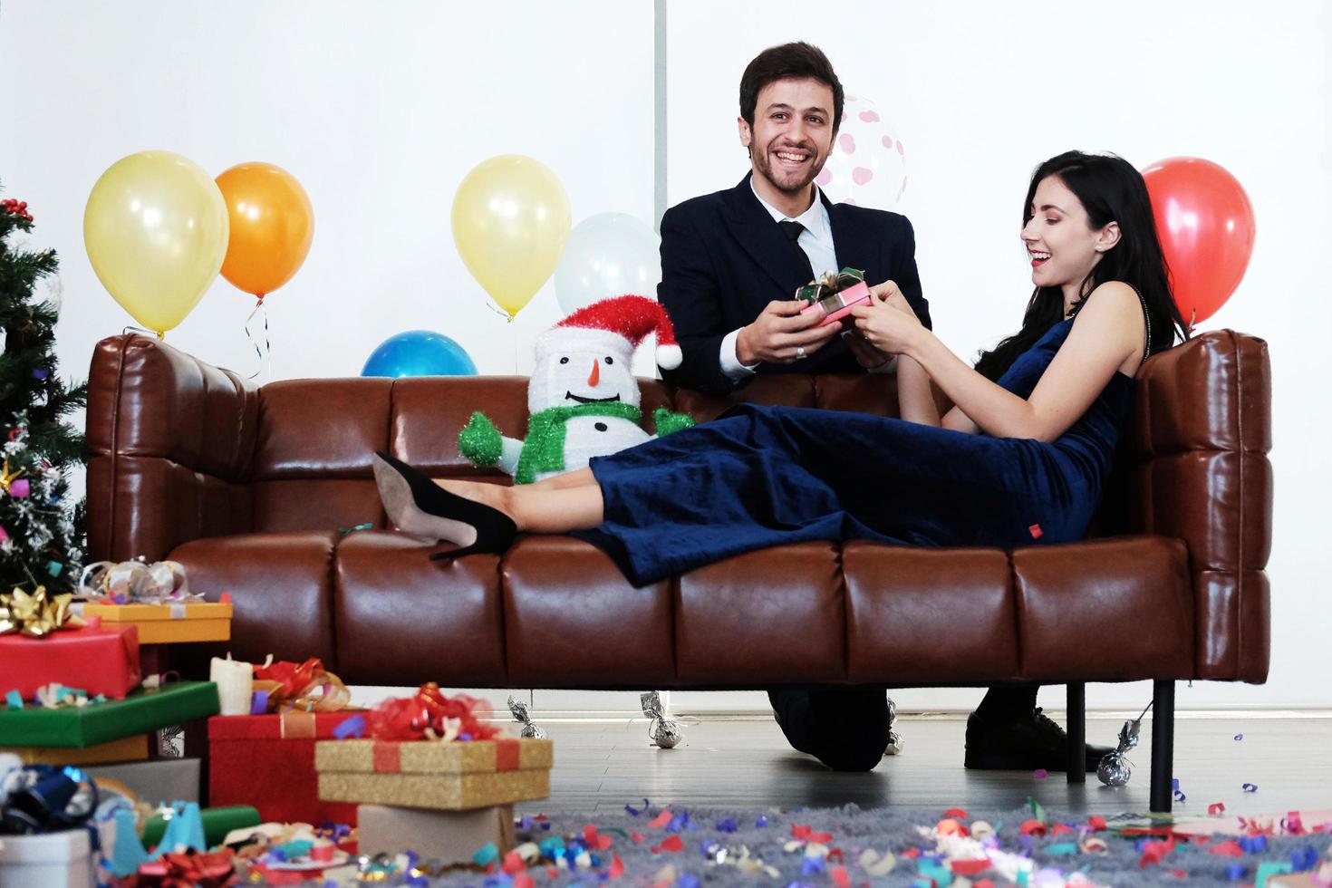 coppia dolce amore felice con scatola regalo a sorpresa fidanzata nel periodo natalizio e festeggia il capodanno, decorazione di San Valentino con albero di Natale, palloncini colorati e scatole regalo sul divano marrone foto