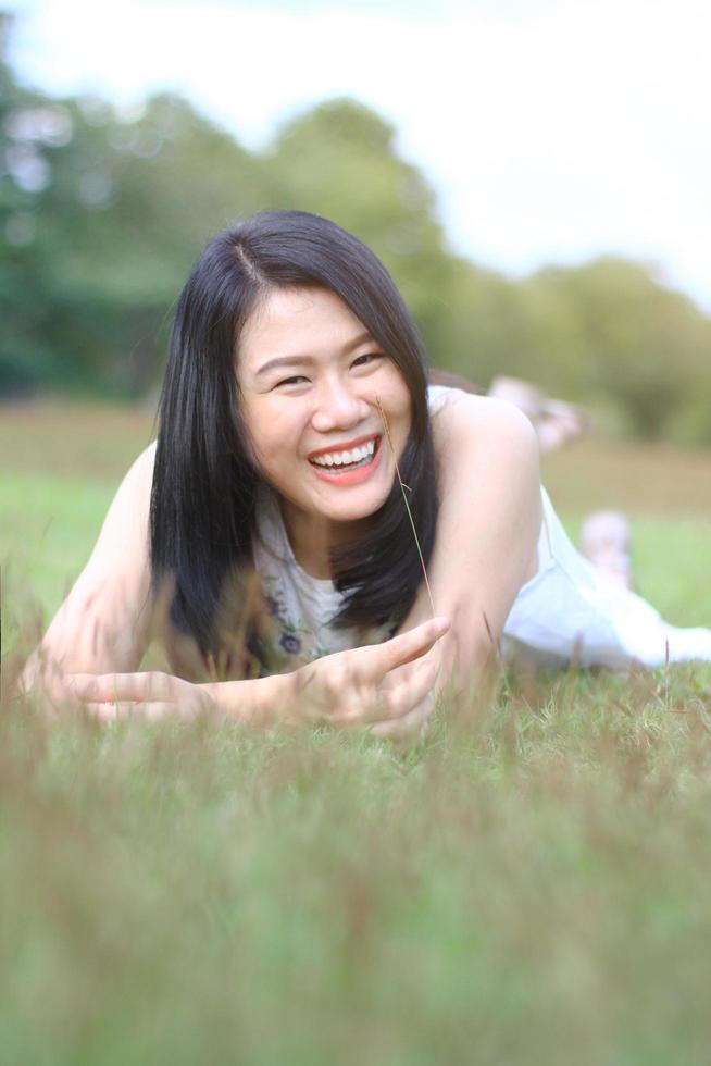 la bella donna asiatica in vestito bianco si rilassa e sorride sul campo verde nel parco naturale. la ragazza tailandese o la ragazza cinese si divertono in vacanza con la luce del sole in giardino foto