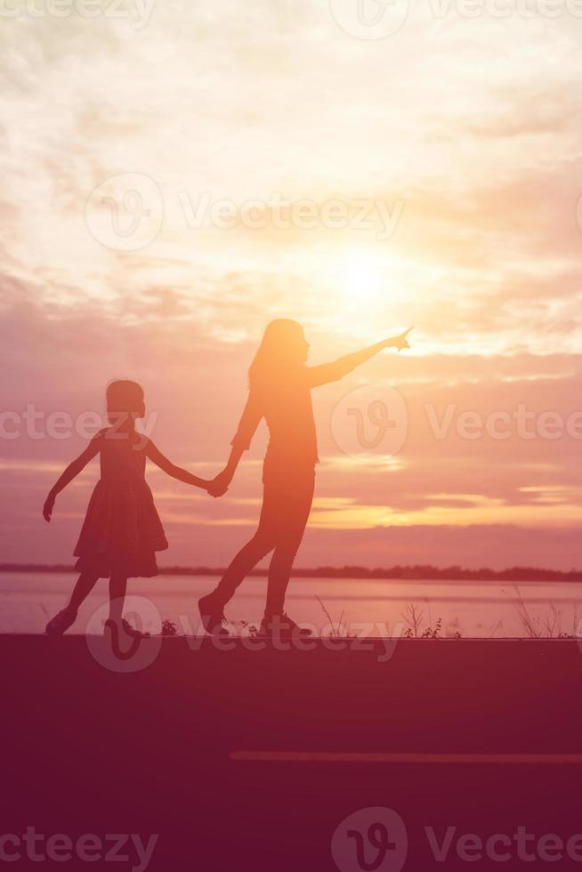 una silhouette di una bambina felice le braccia della sua amorevole madre per un abbraccio, davanti al tramonto nel cielo in una giornata estiva. foto