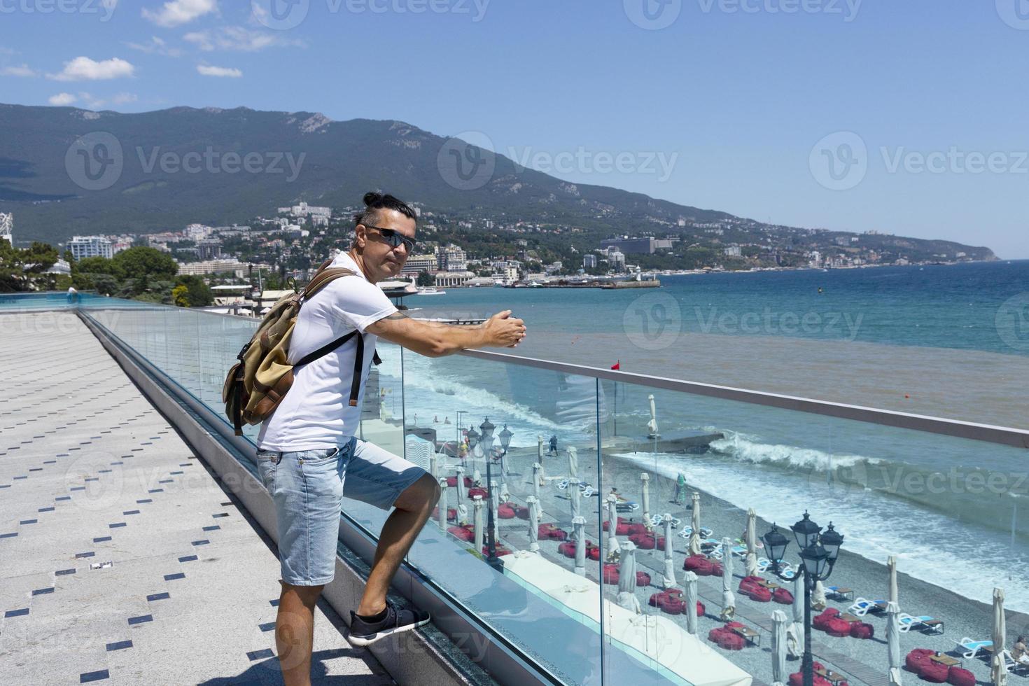 giovane uomo attraente con i capelli scuri in occhiali da sole e uno zaino su un moderno terrapieno in riva al mare in una città europea. vista laterale. foto