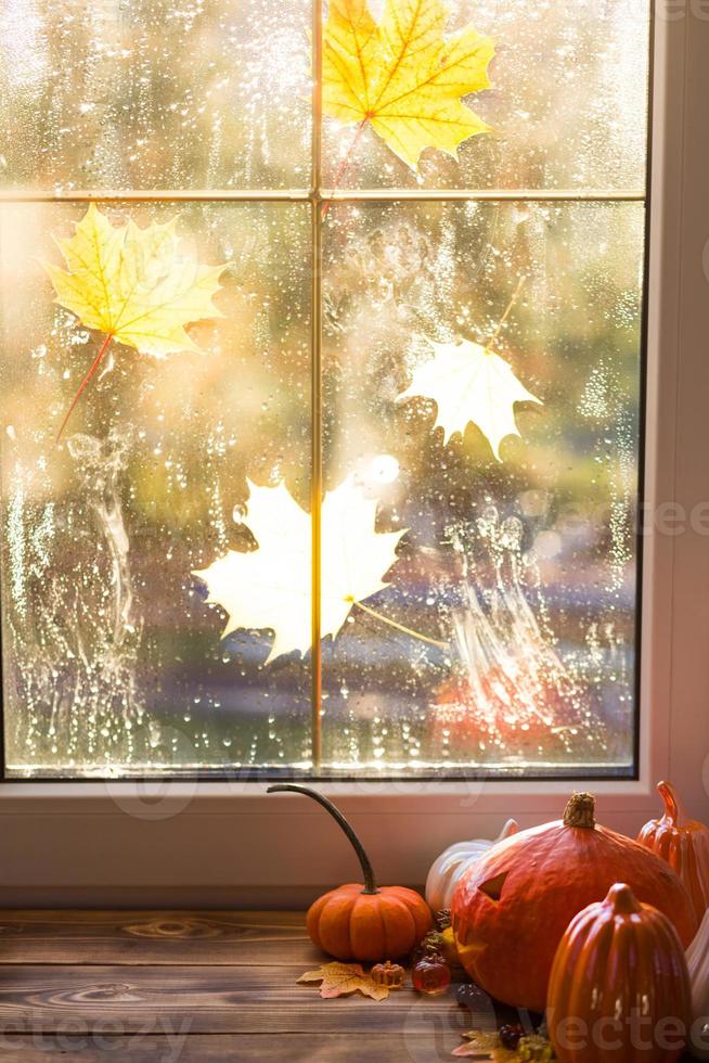 zucca arancione e lanterna jack con gli occhi e la bocca ritagliati dalla finestra con gocce di pioggia su un tavolo di legno con foglie d'acero gialle autunnali. atmosfera autunnale, halloween, arredamento per la casa foto