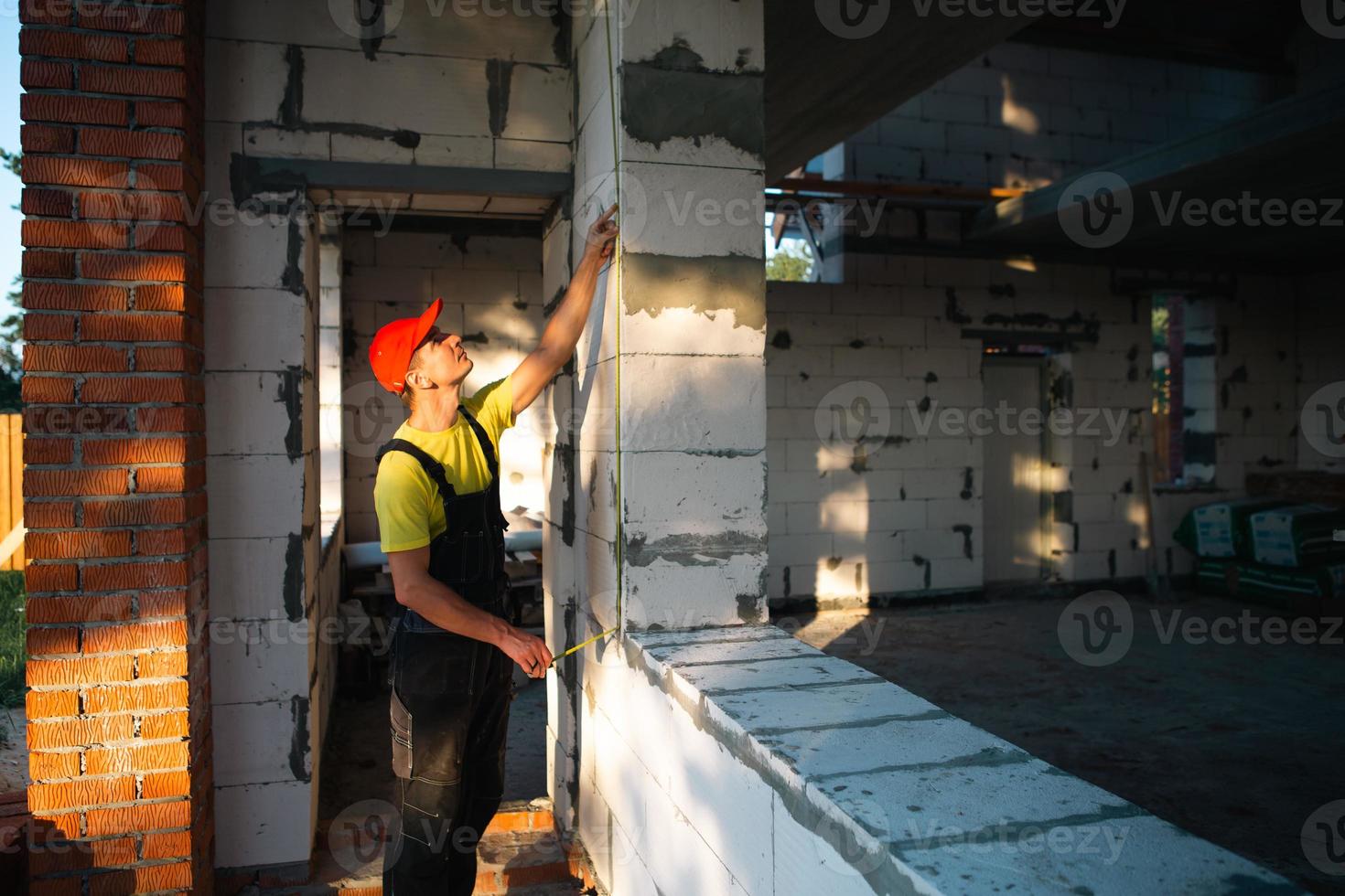 l'operaio edile in cantiere misura la lunghezza dell'apertura della finestra e del muro di mattoni con un metro a nastro. cottage sono fatti di blocchi di cemento poroso, abiti da lavoro - tuta e berretto da baseball foto