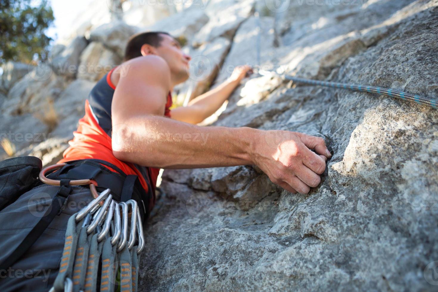 lo scalatore in maglietta rossa si arrampica su una roccia grigia. una mano forte afferrò il comando, messa a fuoco selettiva. forza e resistenza, attrezzatura da arrampicata corda, imbracatura, gesso, sacca portamagnesite, moschettoni, bretelle, rinvii foto