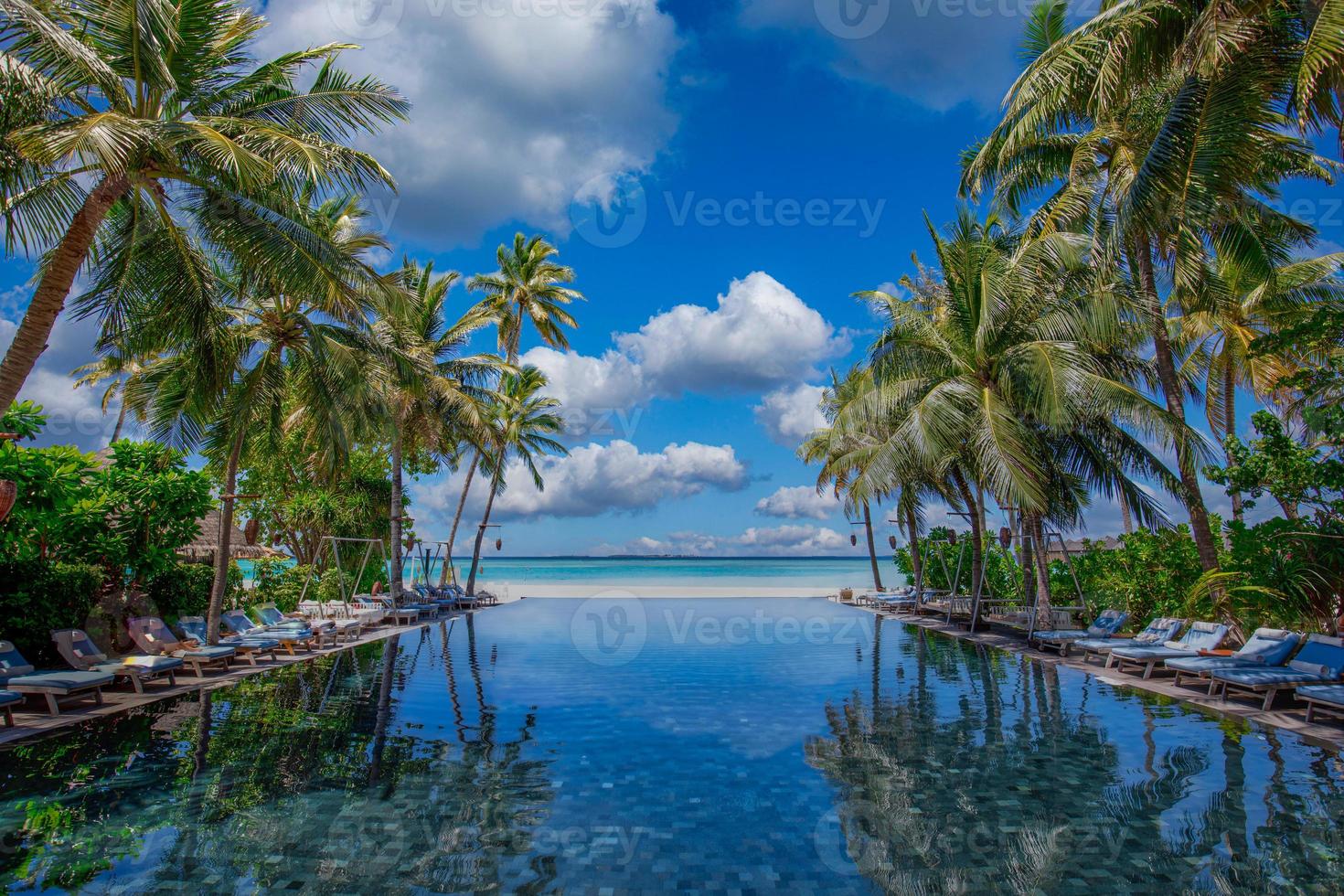 paesaggio del turismo all'aperto. lussuoso resort sulla spiaggia con riflesso sulla piscina, sedie a sdraio o lettini sotto gli ombrelloni con palme e cielo blu. concetto di sfondo di viaggi e vacanze estive foto