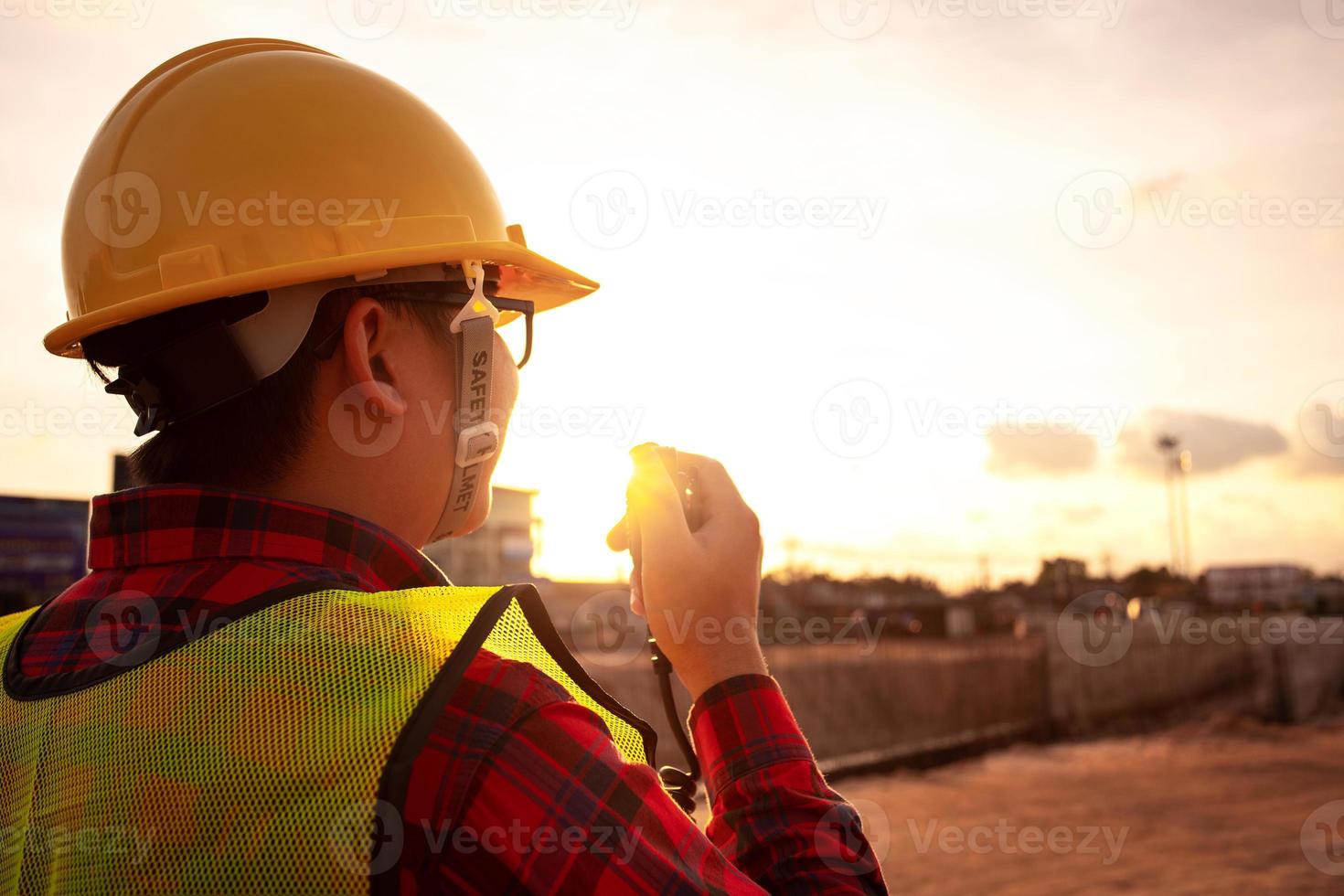 tecnico asiatico ingegnere civile parla con il team di walkie talkie e punta al punto di ispezione per il progetto di lavori di costruzione di trasporto con escavatore sul sito di ingegneria sullo sfondo del tramonto foto