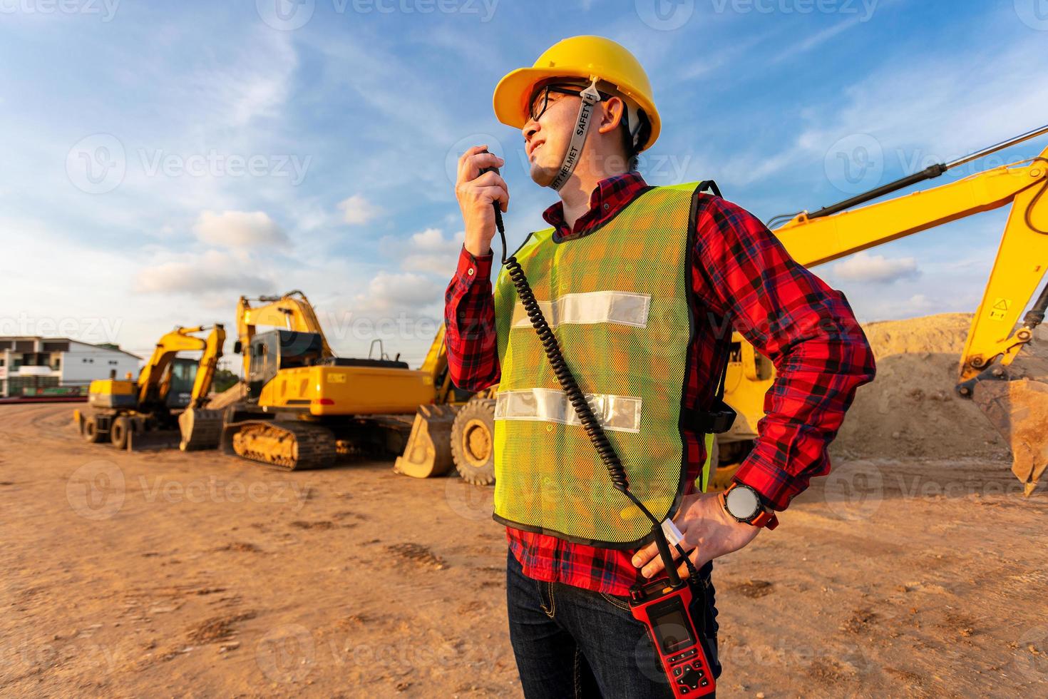 tecnico asiatico ingegnere civile parla con il team di walkie talkie e indica il punto di ispezione per il progetto di lavori di costruzione di trasporto con escavatore sullo sfondo del sito di ingegneria foto