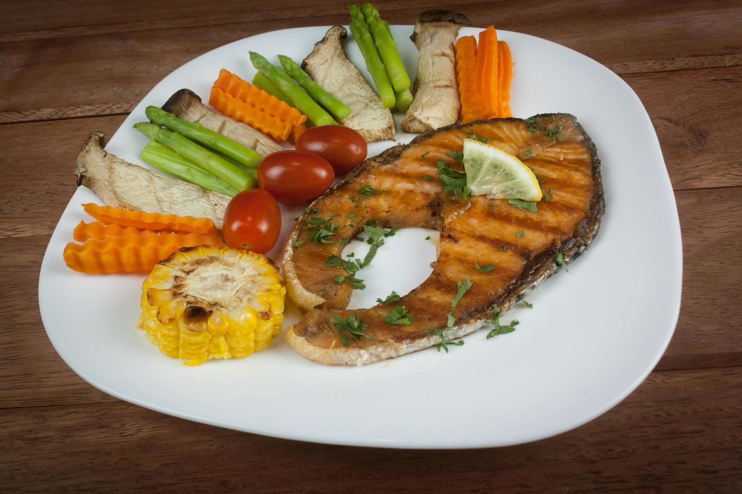 trancio di salmone grigliato scivola fino a cottura, adagiato su un piatto bianco con limone su pesce e verdure, adagiato attorno al piatto su un pavimento di legno. foto