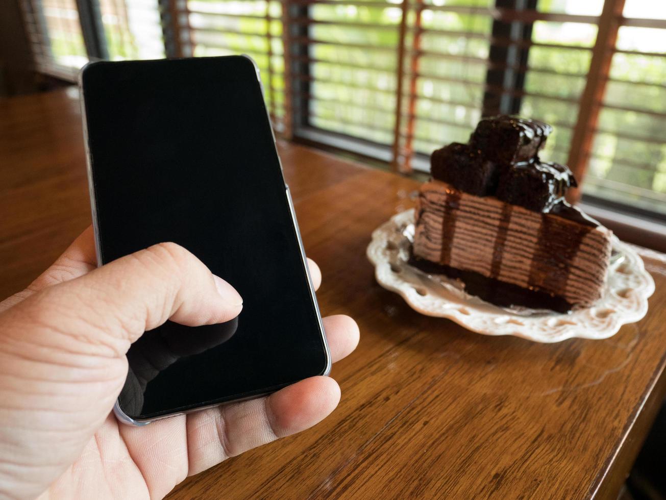 torta al cioccolato con la mano del giovane uomo d'affari che tiene il telefono con lo schermo vuoto per la pubblicità, inserisci un messaggio o un argomento di promozione presso la caffetteria. foto