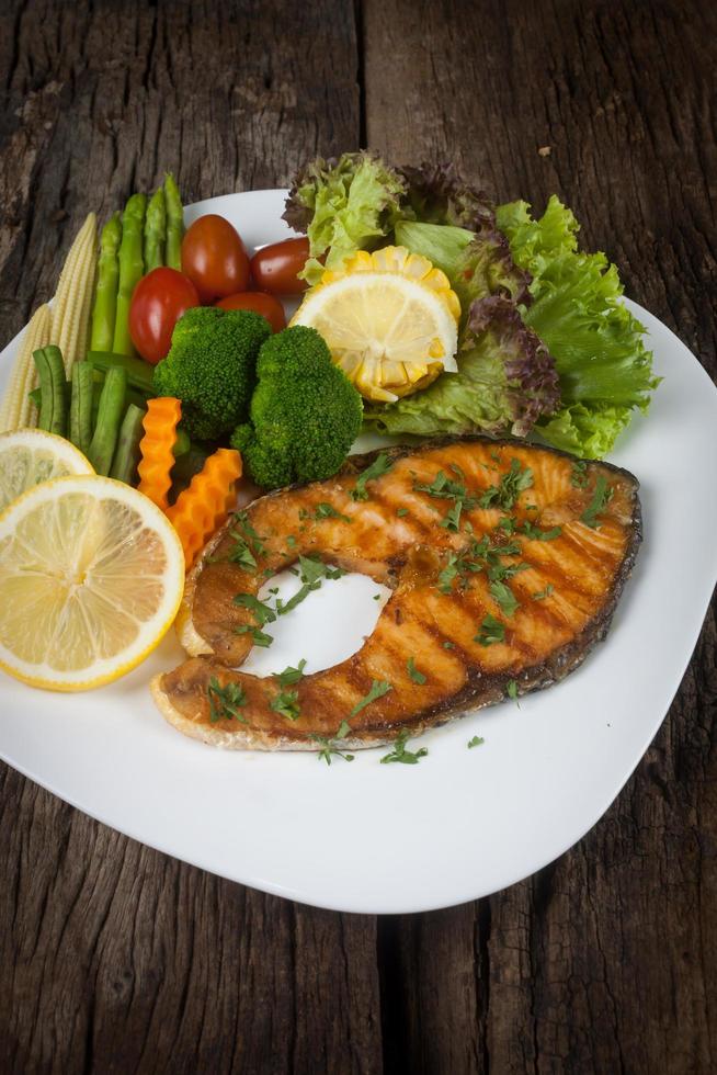 trancio di salmone con salsa su un piatto bianco con limone sul piatto e patatine fritte croccanti. molte verdure sono disposte intorno al piatto sul pavimento di legno. foto