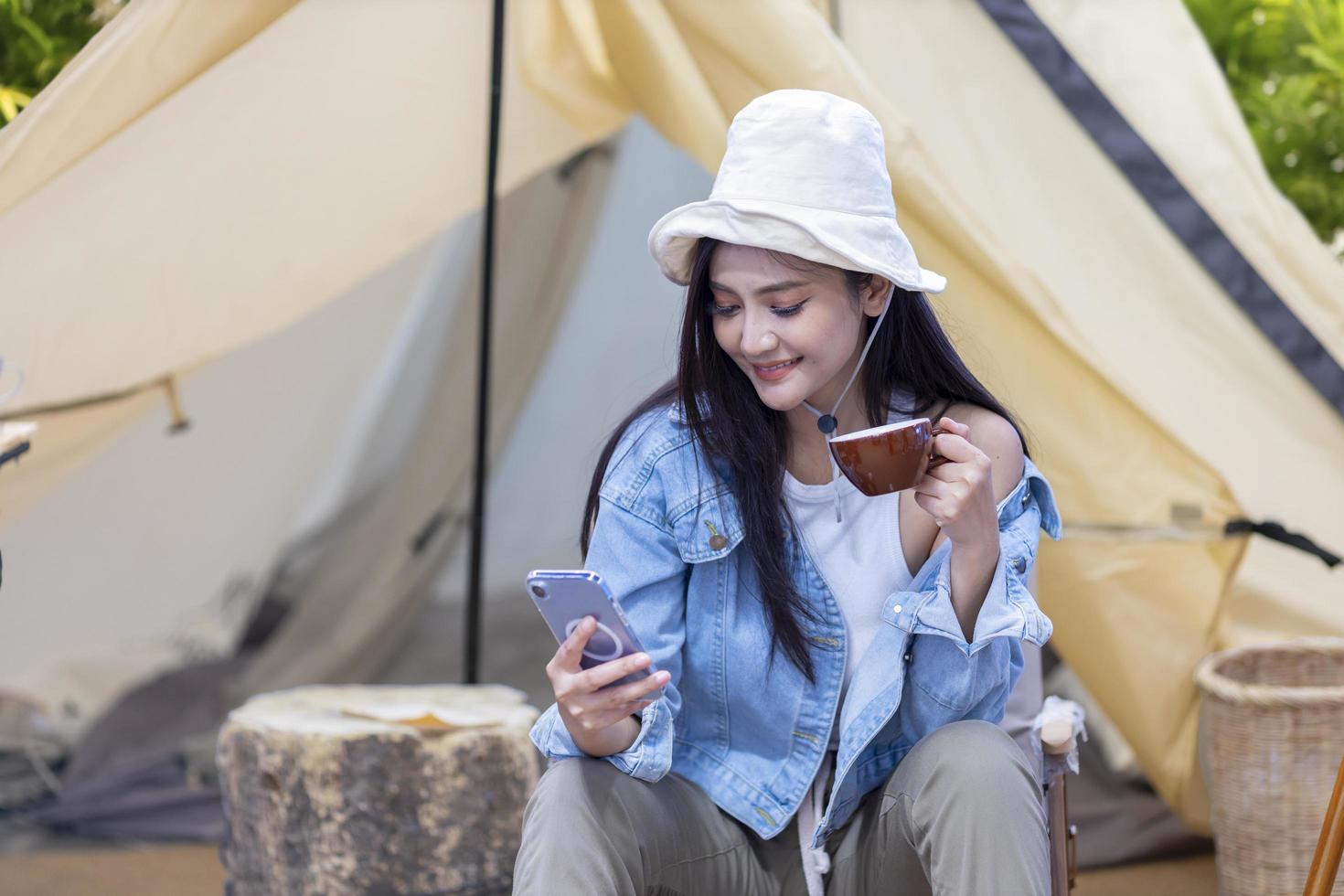 la donna asiatica sta usando il telefono cellulare mentre beve il caffè nella sua tenda mentre si accampa all'aperto durante l'estate nel parco nazionale per l'avventura e il concetto di viaggio attivo foto