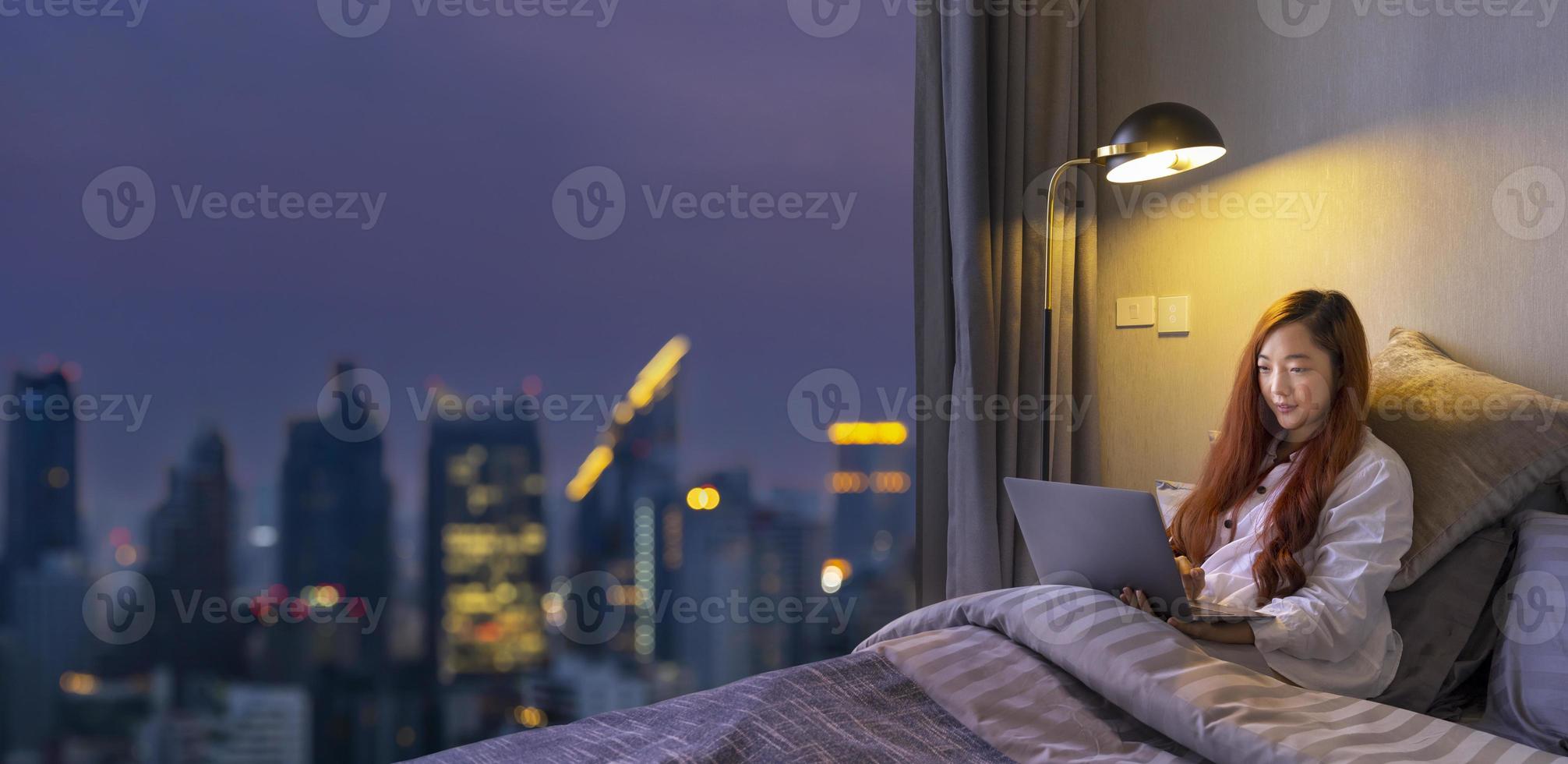 è donna asiatica che lavora online con il laptop nel suo letto a tarda notte con la vista sulla città dell'edificio del grattacielo per lo stile di vita di lusso urbano e il concetto di istruzione foto