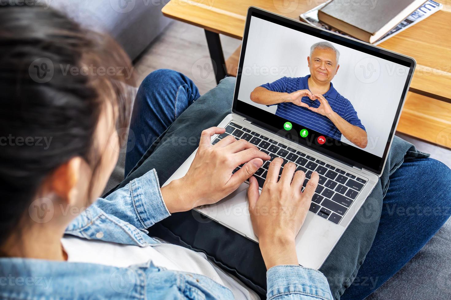 vista posteriore della ragazza asiatica riposa a casa parla in videochiamata sul laptop con felice padre anziano, giovane donna si rilassa sul divano parla chat online con sorridente papà maturo, usa la webcam sul computer foto