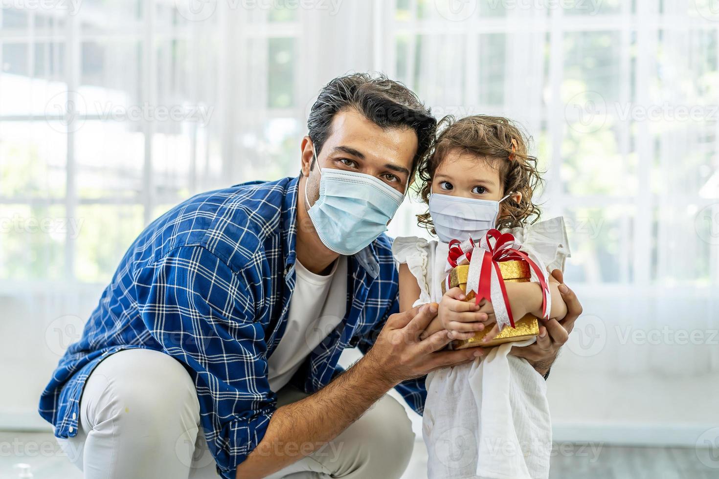 felice padre e figlia in maschera medica con confezione regalo. celebrazione delle vacanze online di natale, natale, capodanno in quarantena coronavirus covid-19 di blocco. nuova normalità, distanza sociale, stare a casa foto