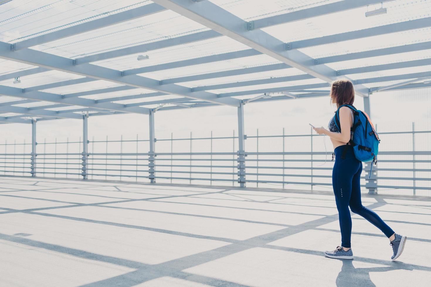 vista posteriore di una giovane donna sportiva in abiti sportivi e scarpe da ginnastica, porta lo zaino, usa un telefono cellulare aggiornato, cammina da qualche parte, ha uno stile di vita sano, gode del tempo libero e di internet ad alta velocità foto